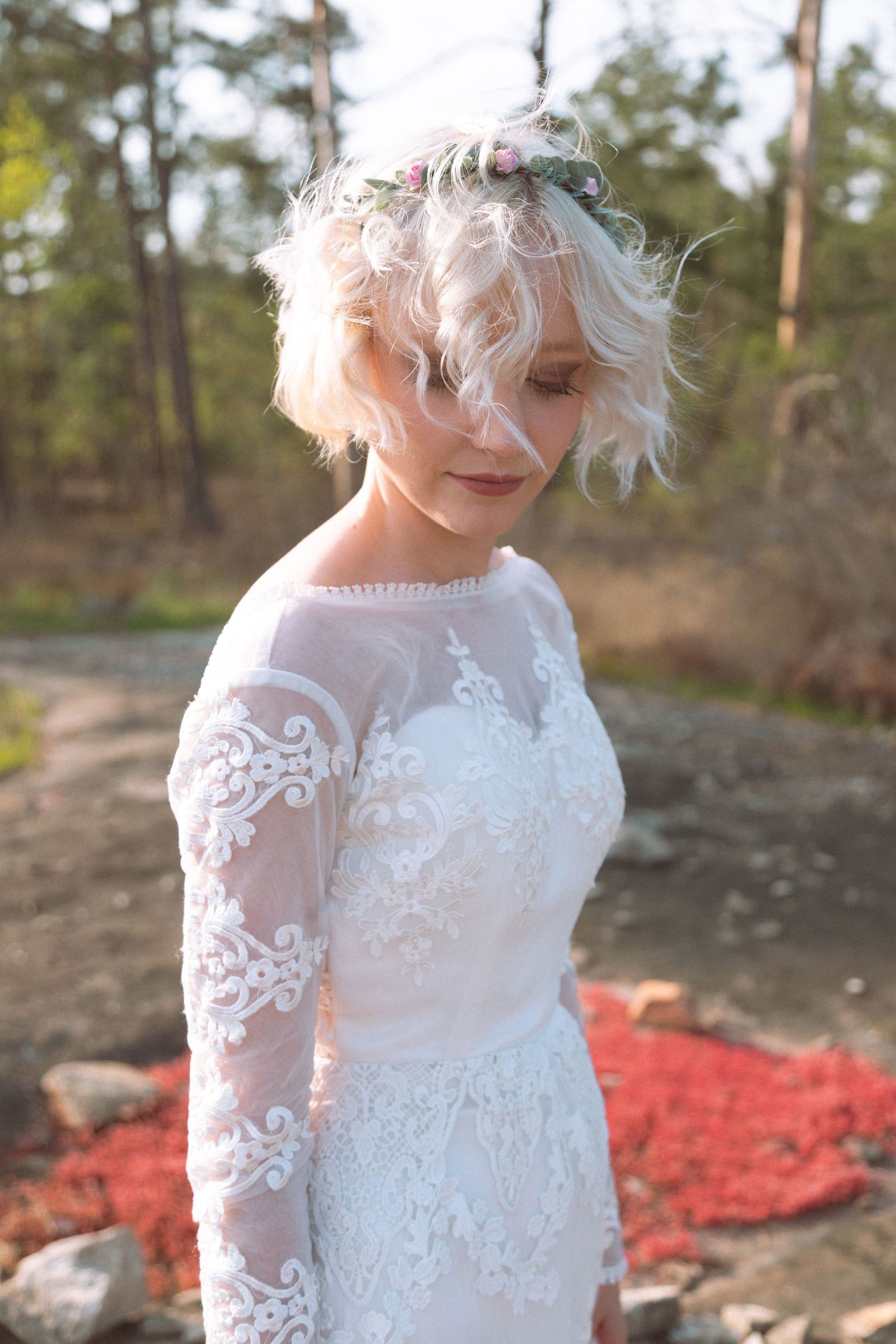 Bride in lace wedding gown with floral crown poses in sunlit forest setting.