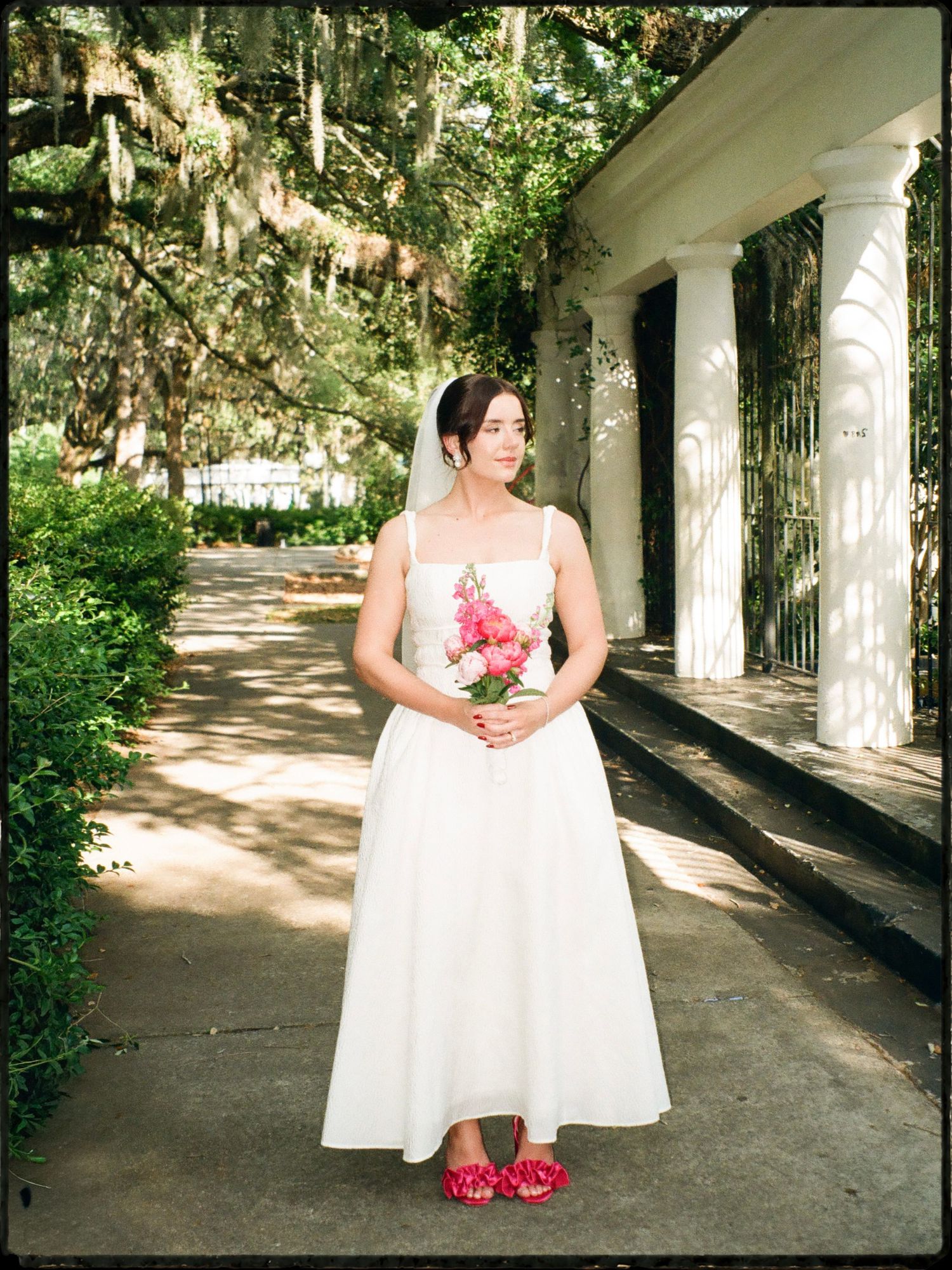 Bride in white sleeveless gown with red flats holds pink bouquet beside columned building outdoors.