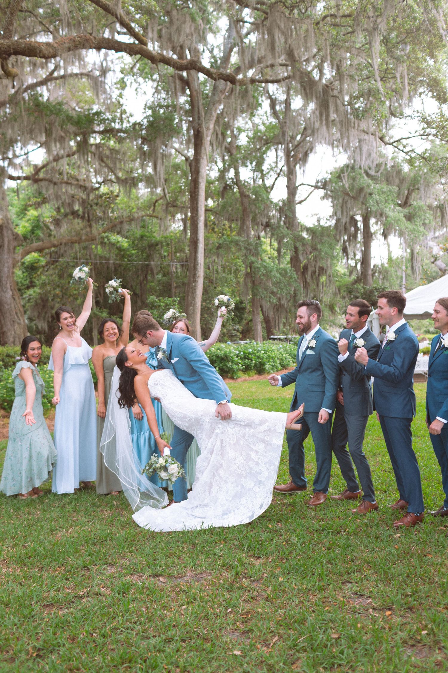 Wedding party celebrates as groom dips bride for a kiss on lush green lawn surrounded by moss-draped oak trees.