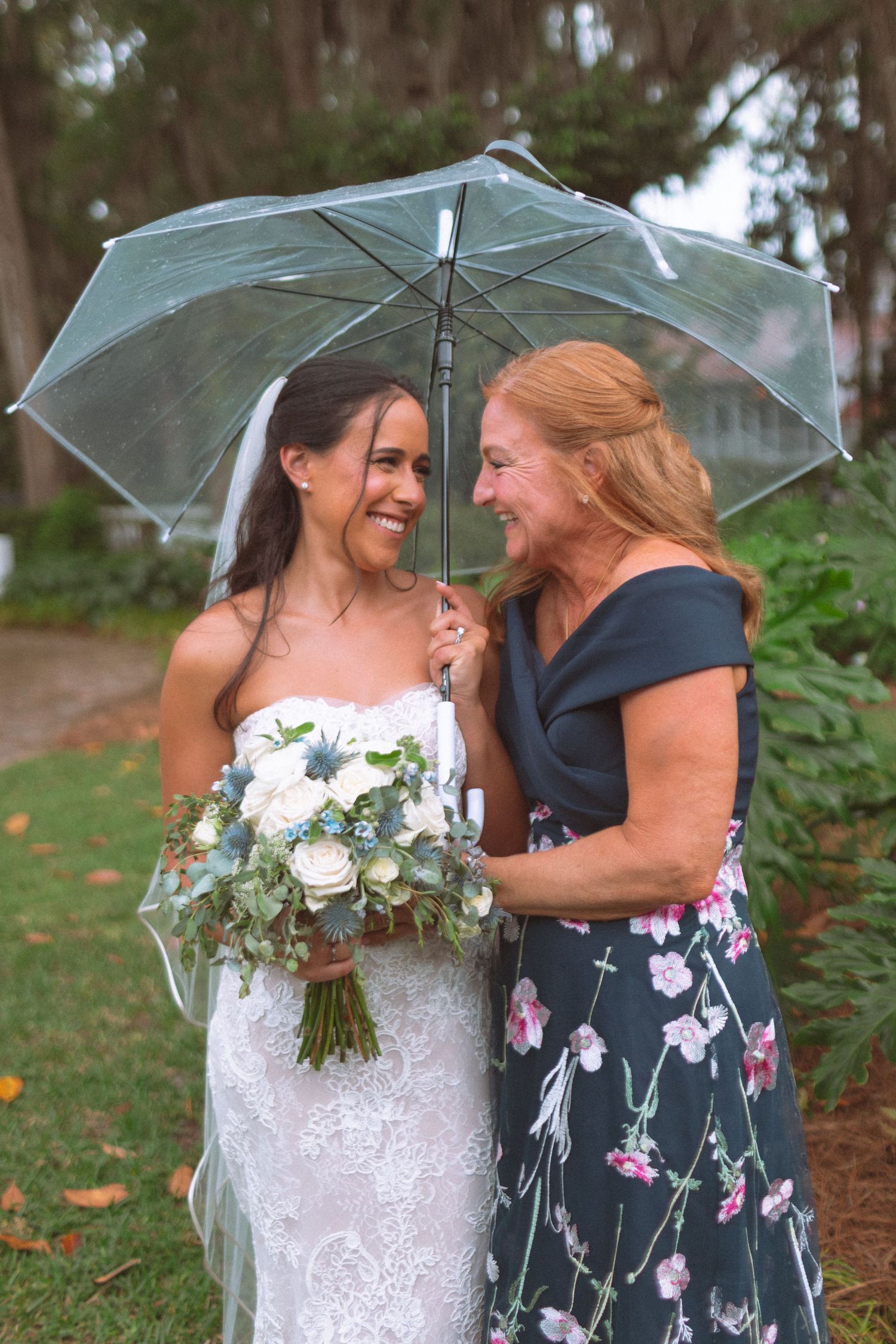 Smiling bride with bouquet shares a joyful moment with her mother under a clear umbrella in the rain.