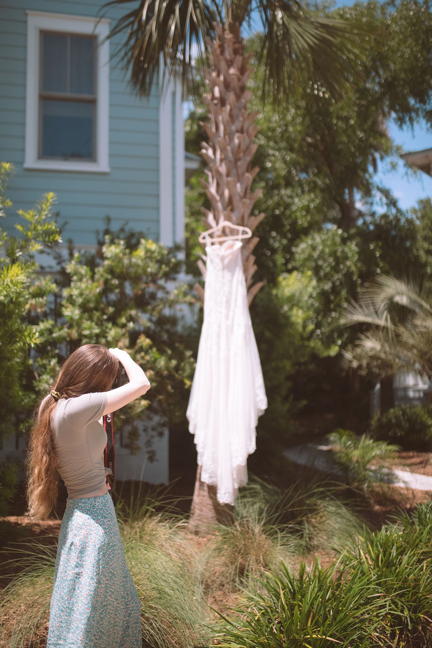 A bride with long hair admires her white wedding dress hanging on a palm tree in a lush garden.