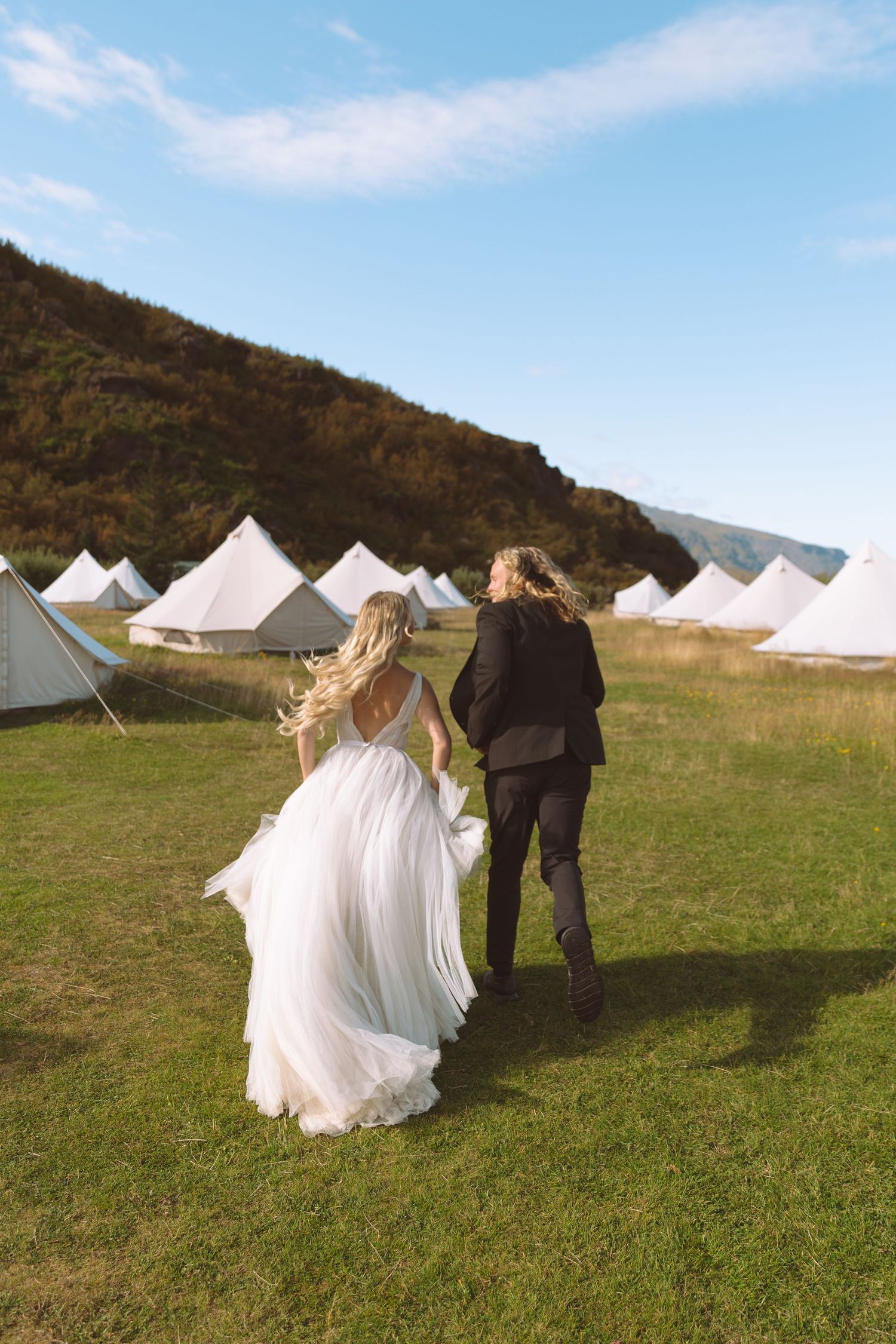 Bride and groom walking hand in hand through a green field with white bell tents and mountains in the background.