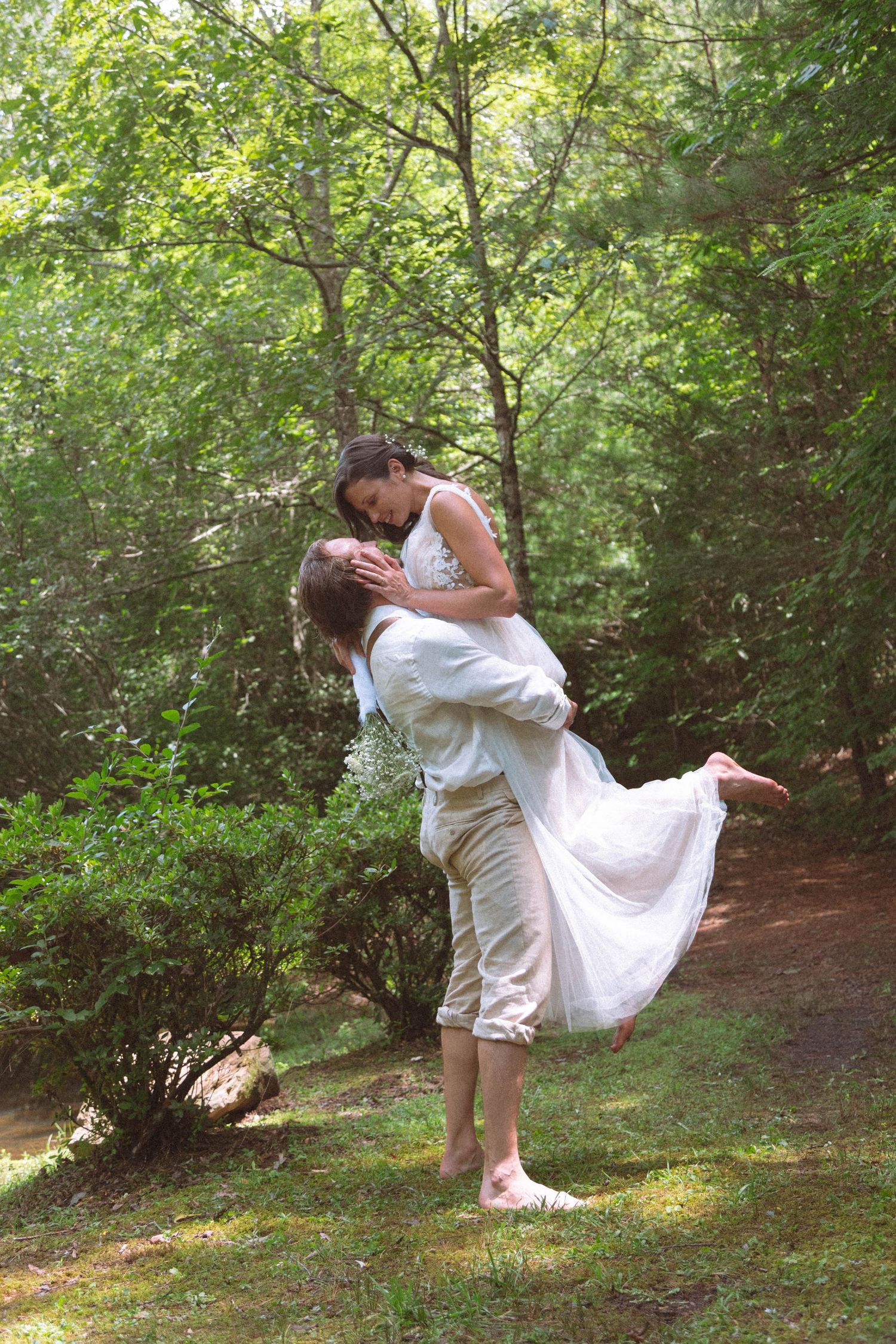 Groom lifting bride in white dress in a sunlit forest path barefoot.