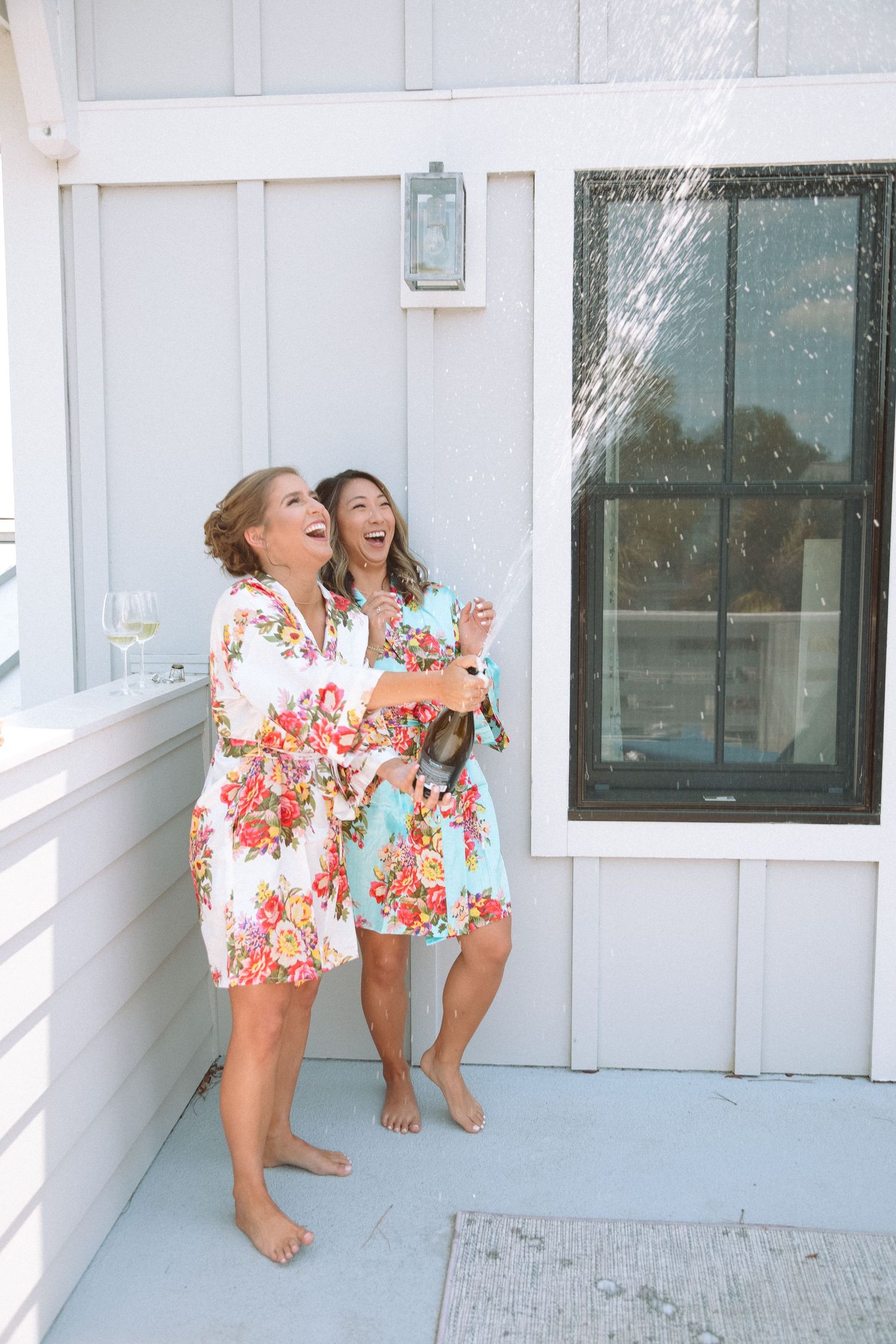 Two bridesmaids in floral robes laughing while popping champagne on a white porch.