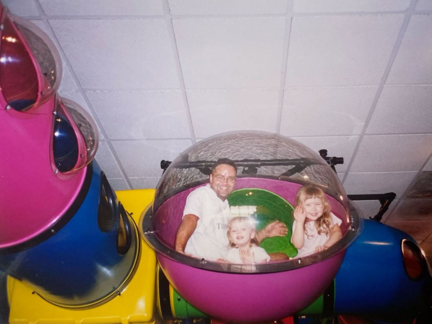 Father and two children sitting inside a colorful dome-shaped ride attraction at an indoor amusement center.
