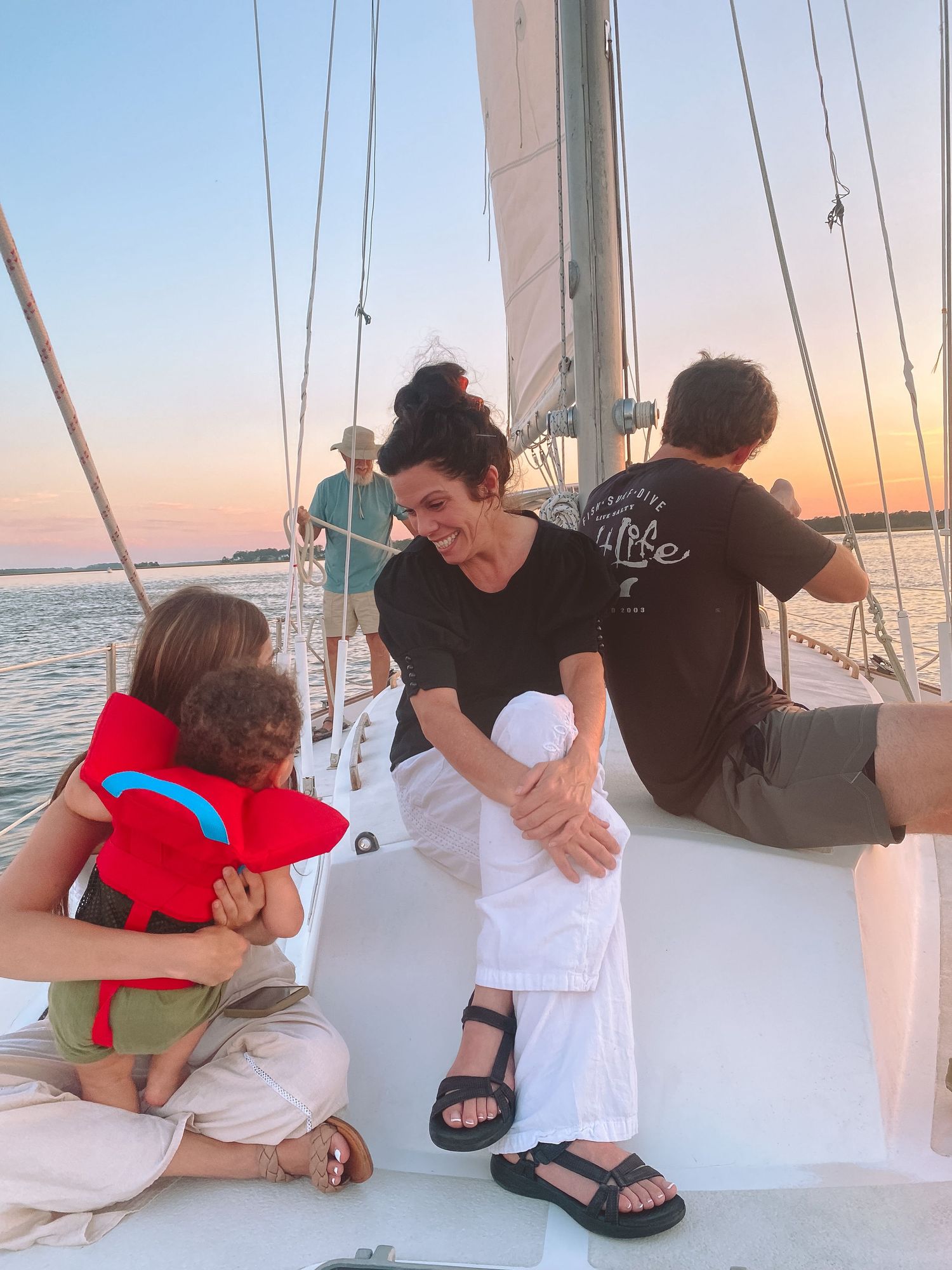 Family relaxing on sailboat deck at sunset, child in red life jacket with adults enjoying the view.