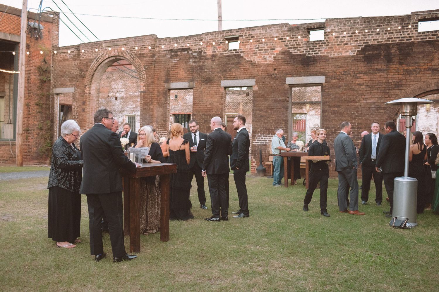 Wedding cocktail hour guests mingling outdoors in a rustic brick ruins venue with string lights overhead.