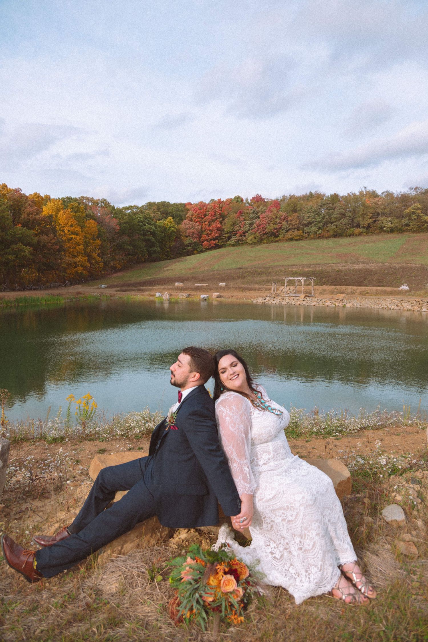 Bride and groom sitting back to back on rocks beside scenic autumn lake with fall foliage.