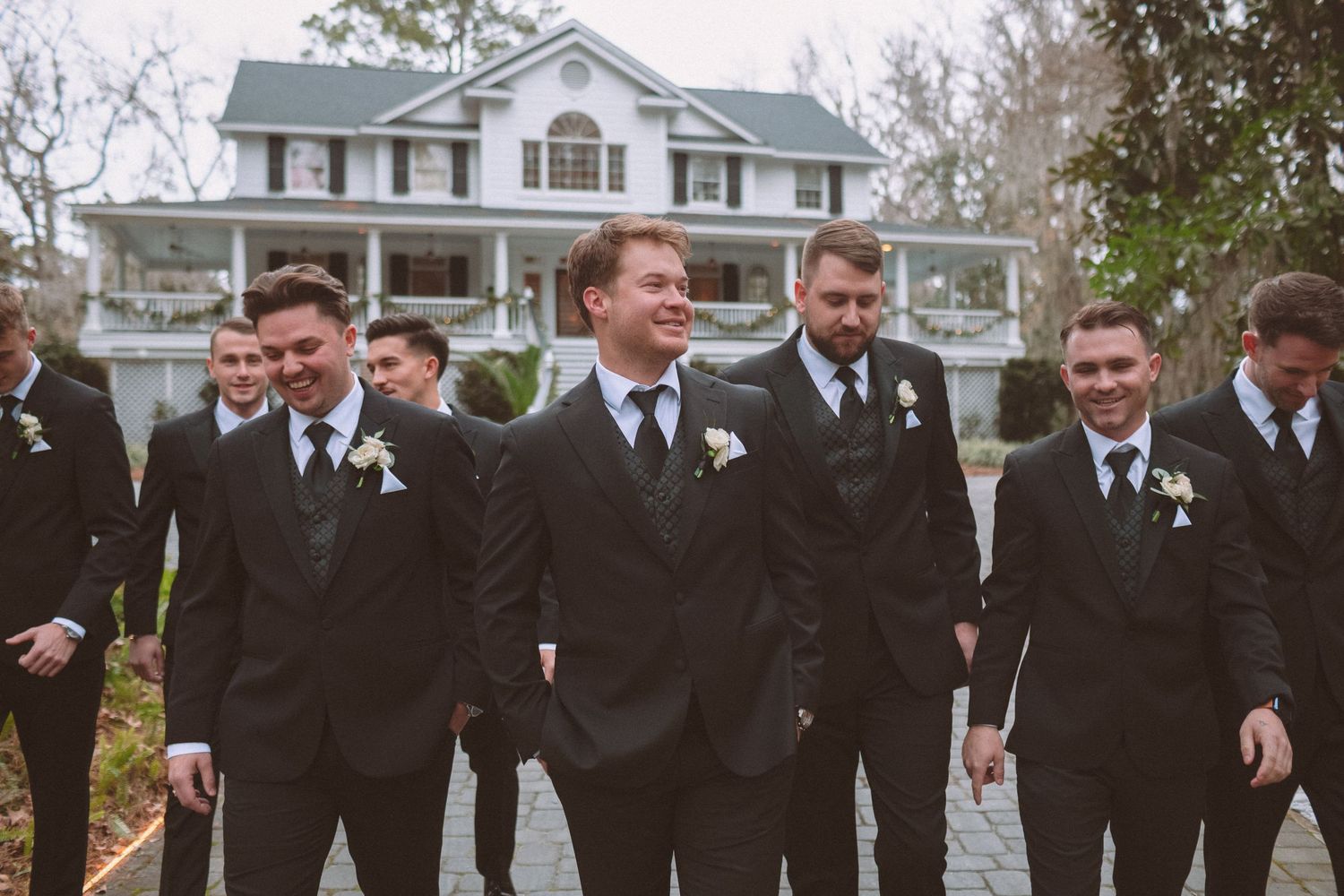 Groom and groomsmen in black suits walk together laughing in front of a white manor house.