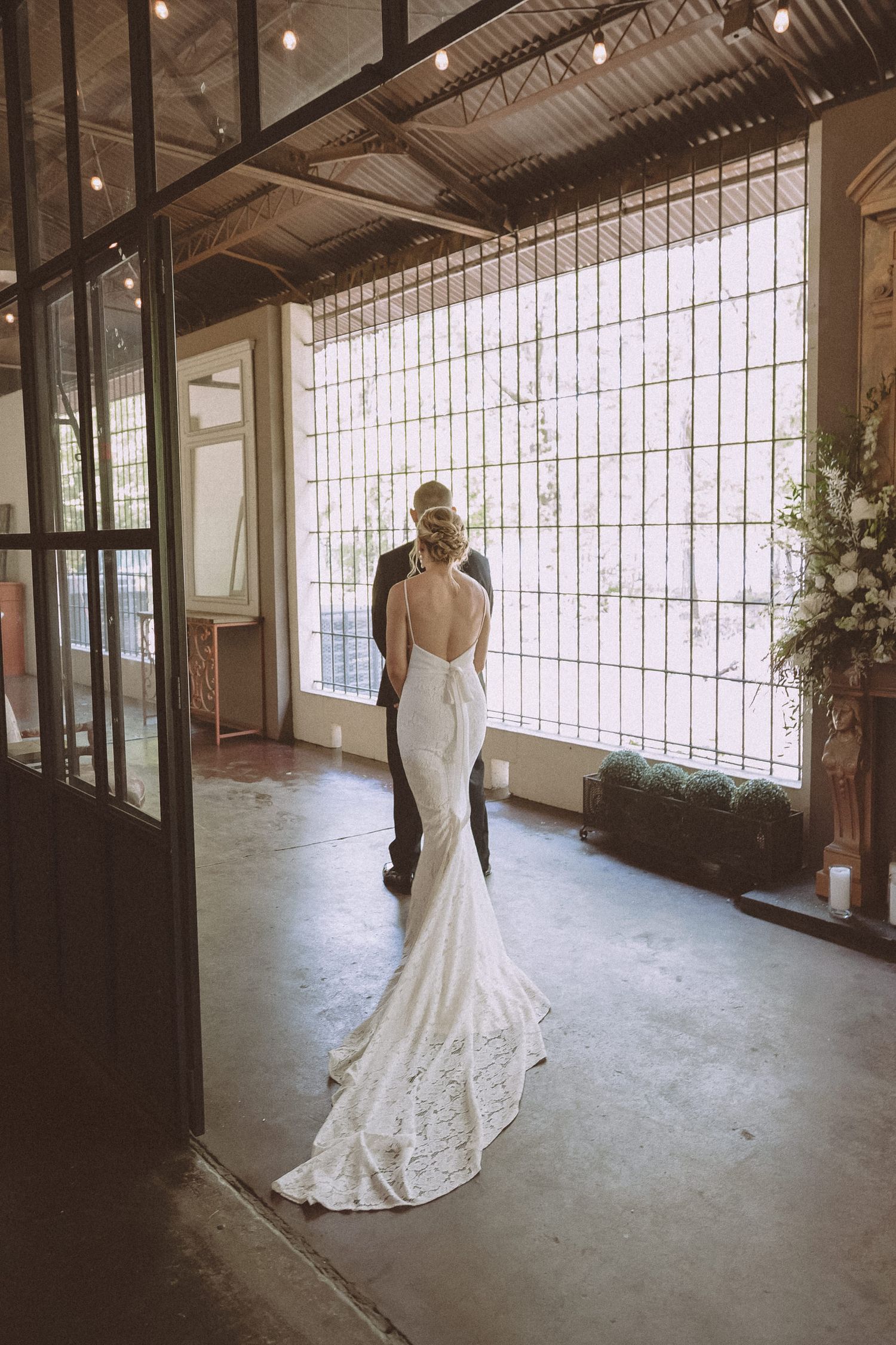 Bride in elegant backless lace gown with train stands with groom near large industrial windows.
