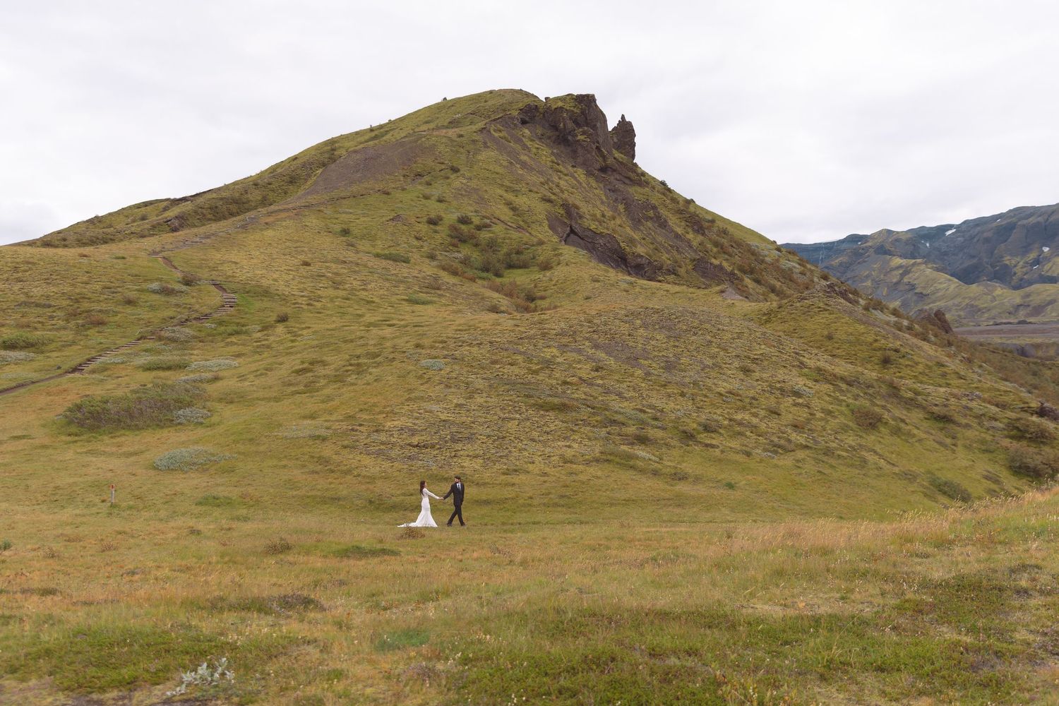 Bride and groom walk hand in hand across sweeping green mountain valley during outdoor wedding shoot.