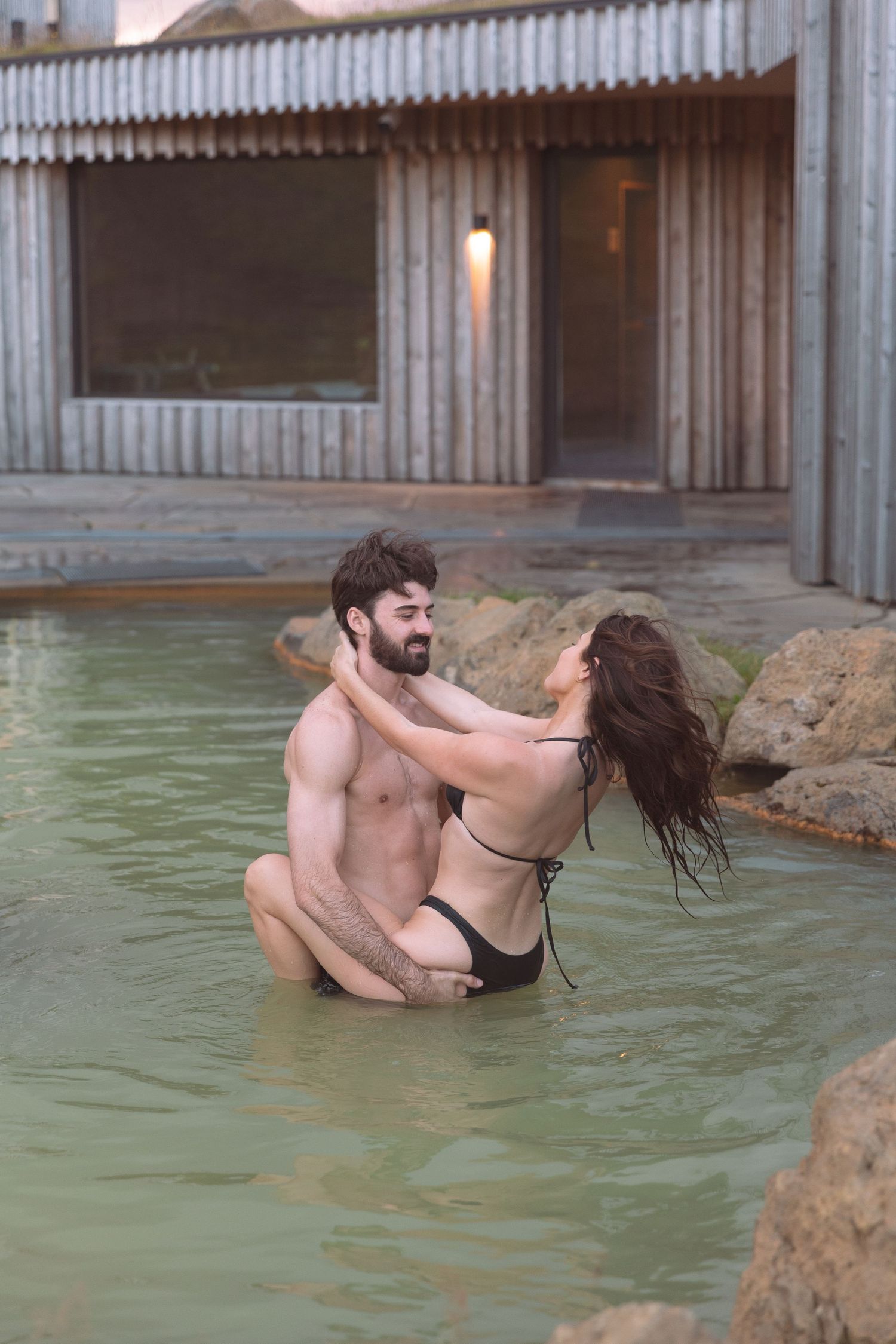 Romantic couple embracing in a geothermal hot spring pool near a wooden spa building.