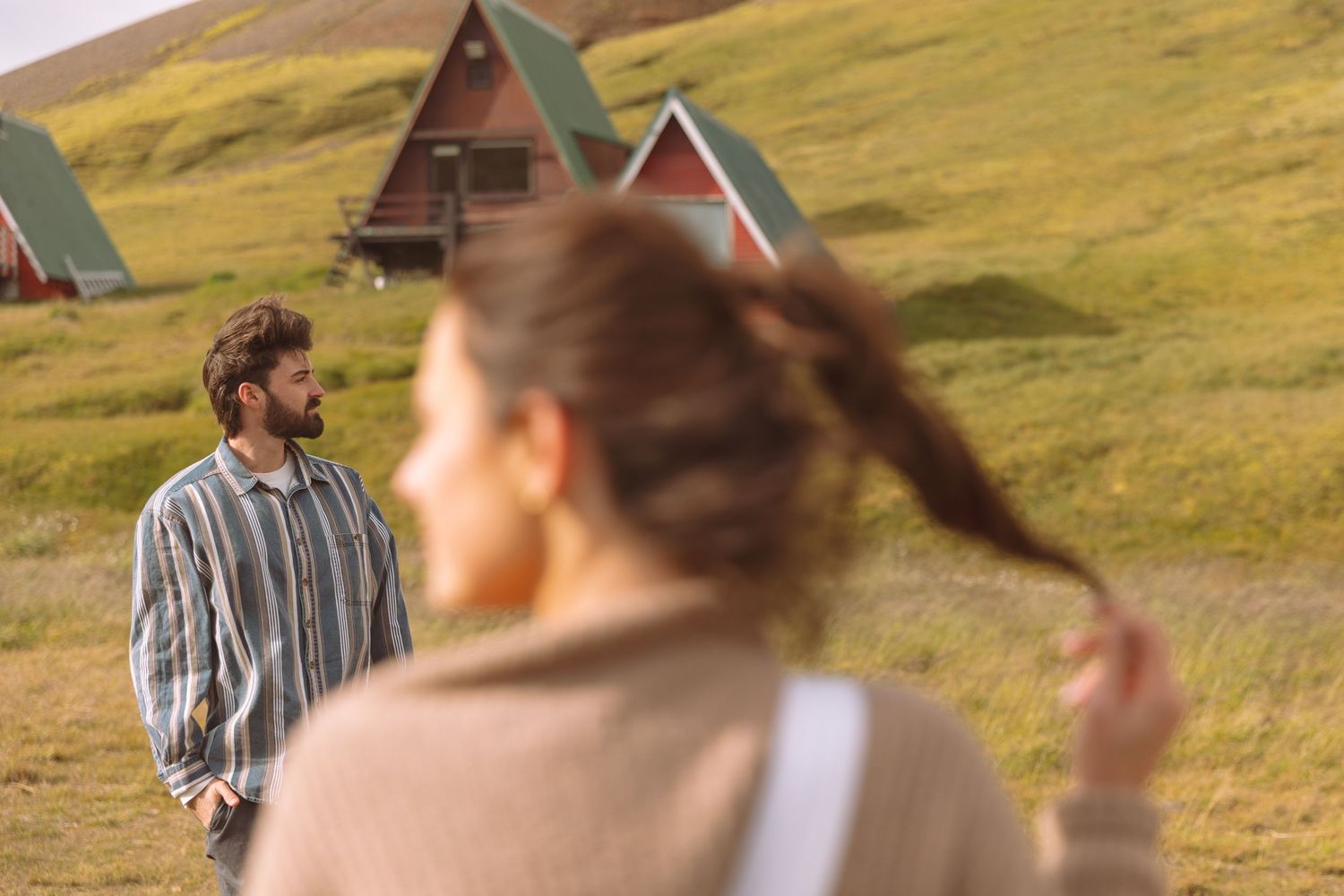 A man and woman stand apart in a green meadow with red-roofed A-frame cabins in the background.