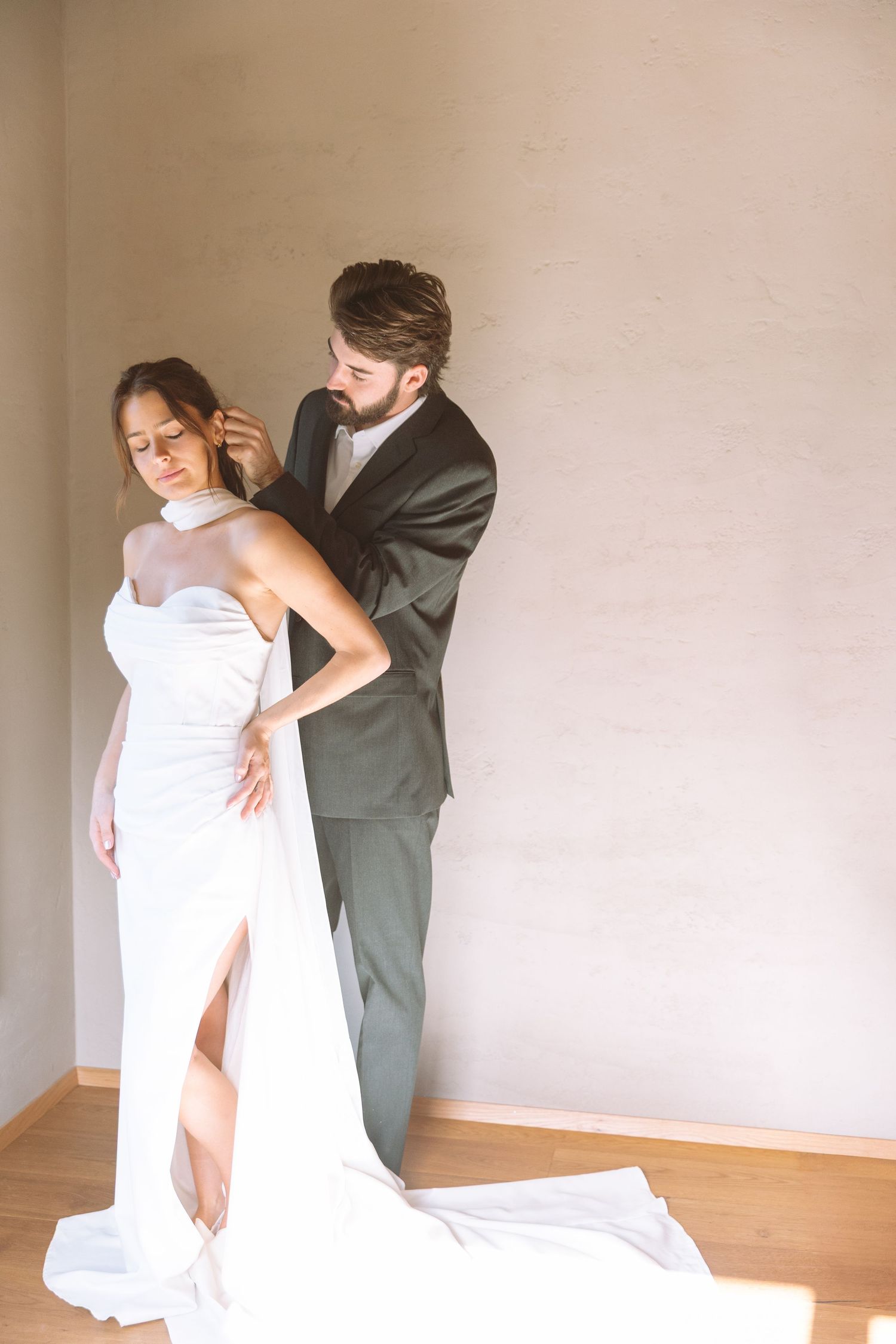 Groom fastening brides halter neck wedding dress against a soft neutral wall.