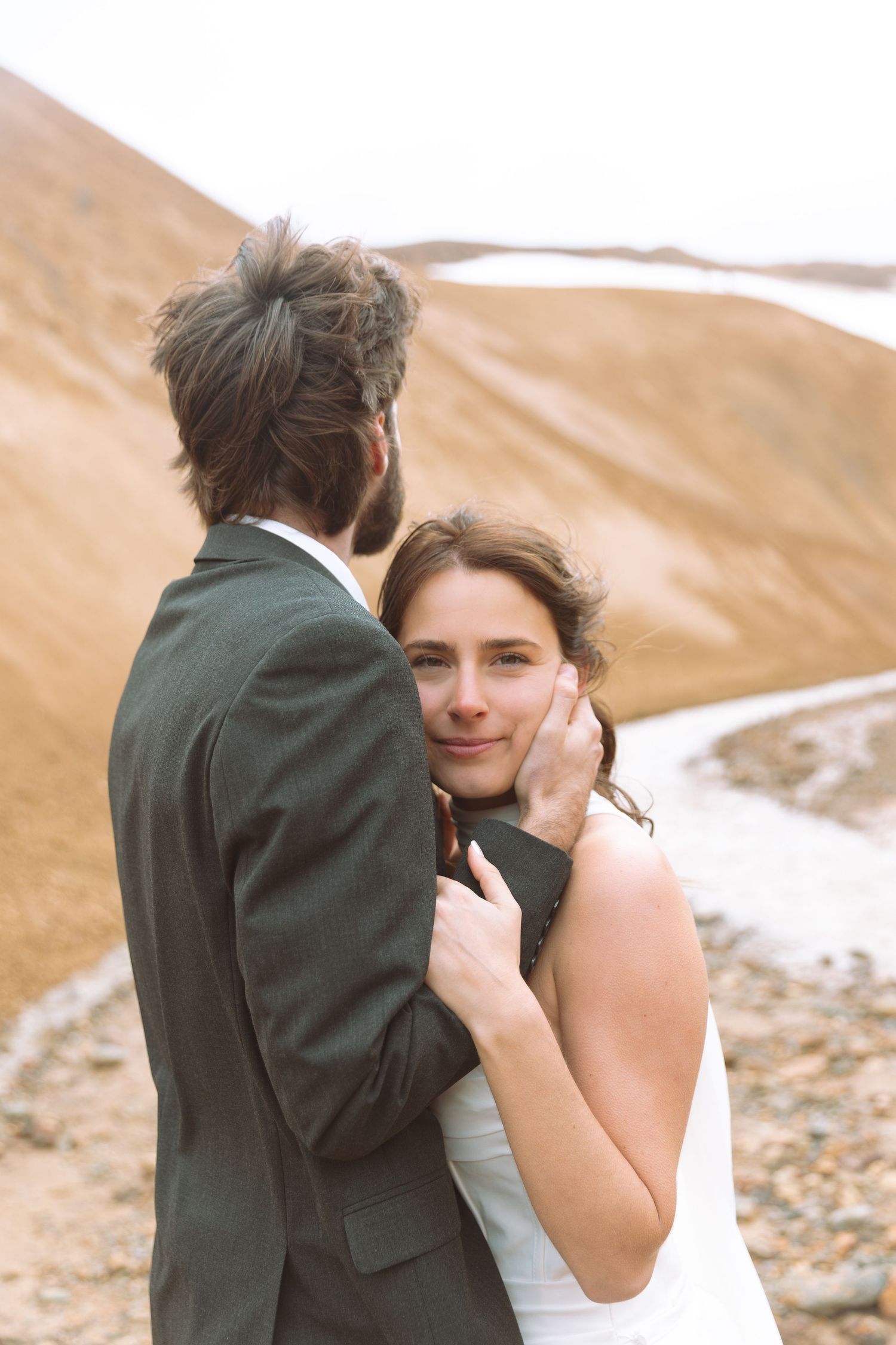 Smiling bride in white dress embraces groom from behind, looking at camera with sandy hills behind them.