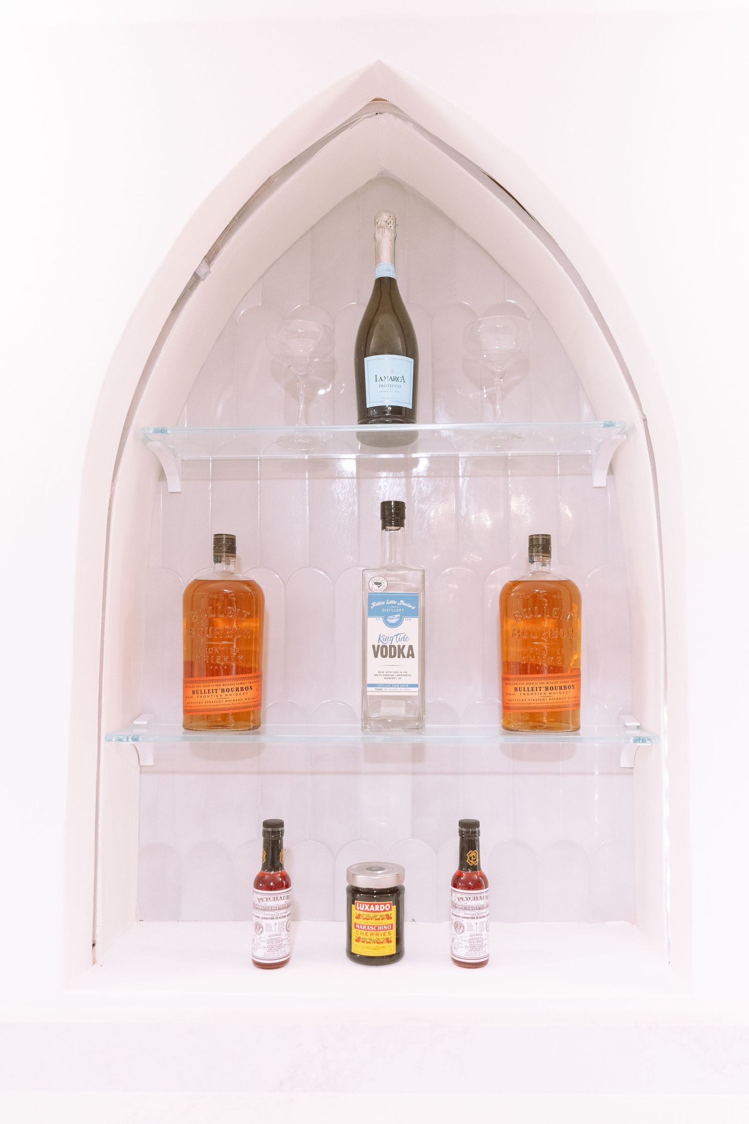 Gothic arch bar niche with vodka, bourbon bottles, bitters, and champagne on glass shelves.