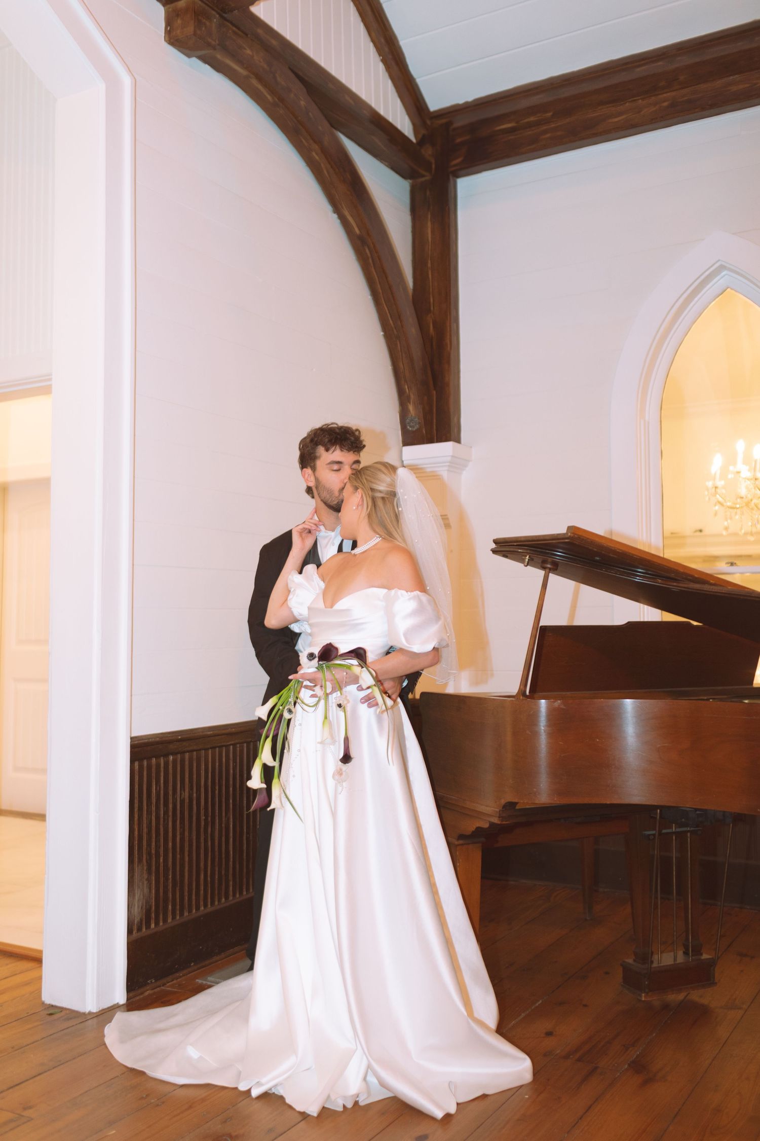 Bride and groom sharing a kiss beside a grand piano in an elegant chapel venue.