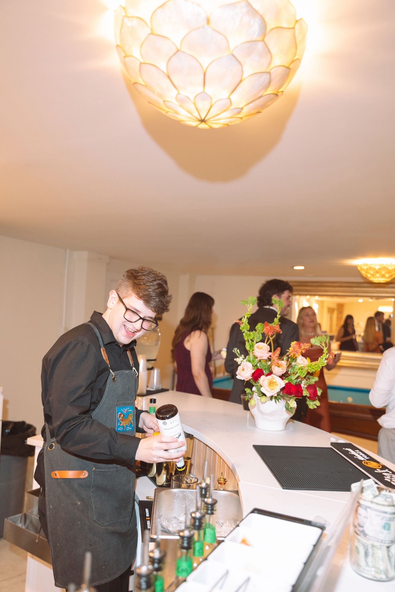 Young bartender in black apron preparing drinks at elegant event bar with floral arrangement and chandelier.