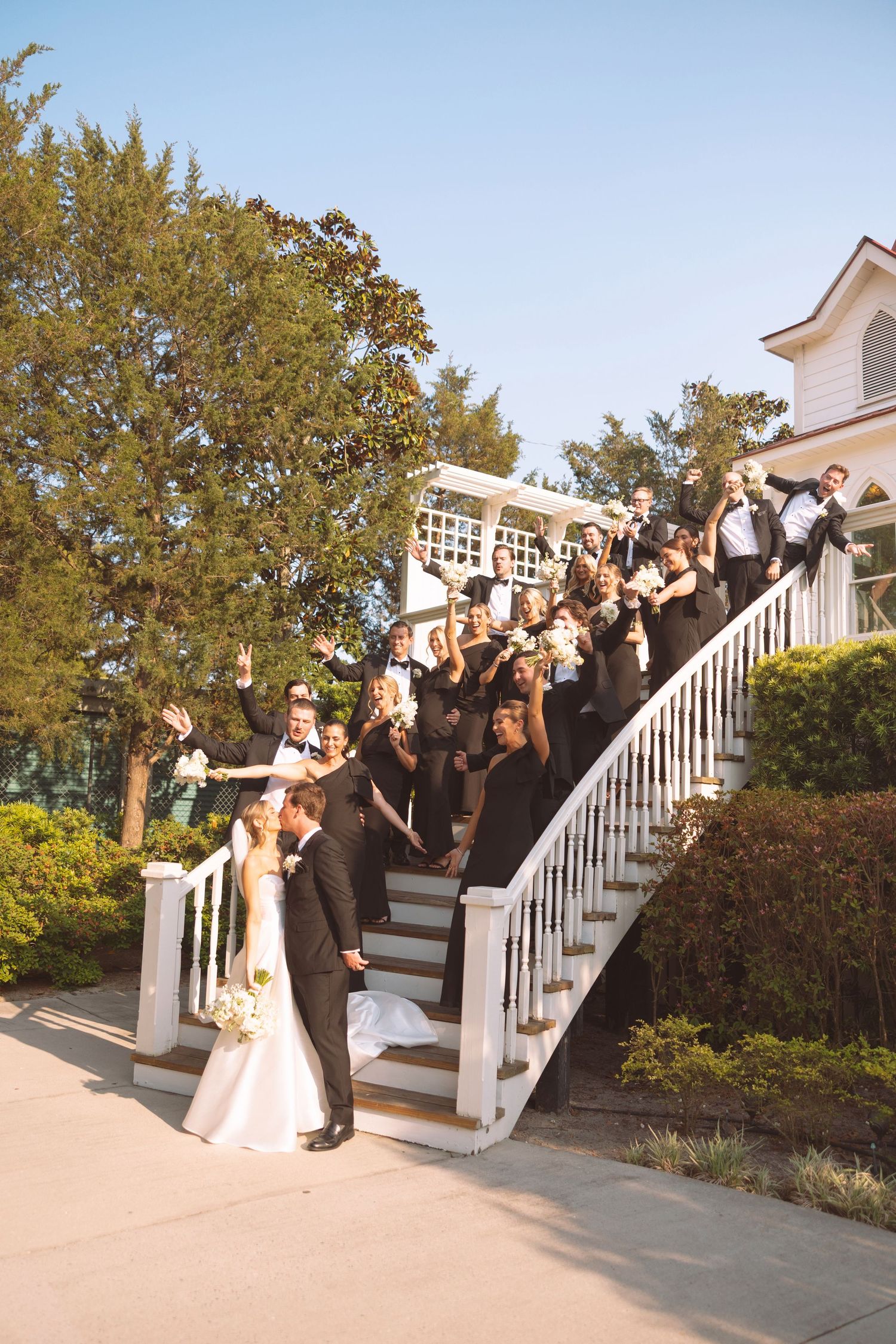 Wedding party in black attire cheering on white curved staircase outside elegant white mansion with bride and groom kissing.