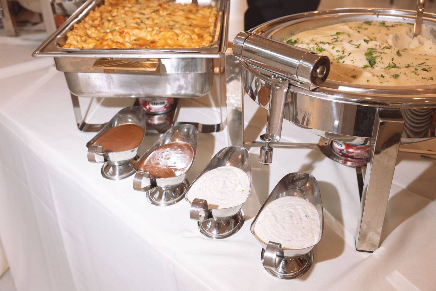 Row of silver chafing dishes with ladles on a white tablecloth at a catered event.