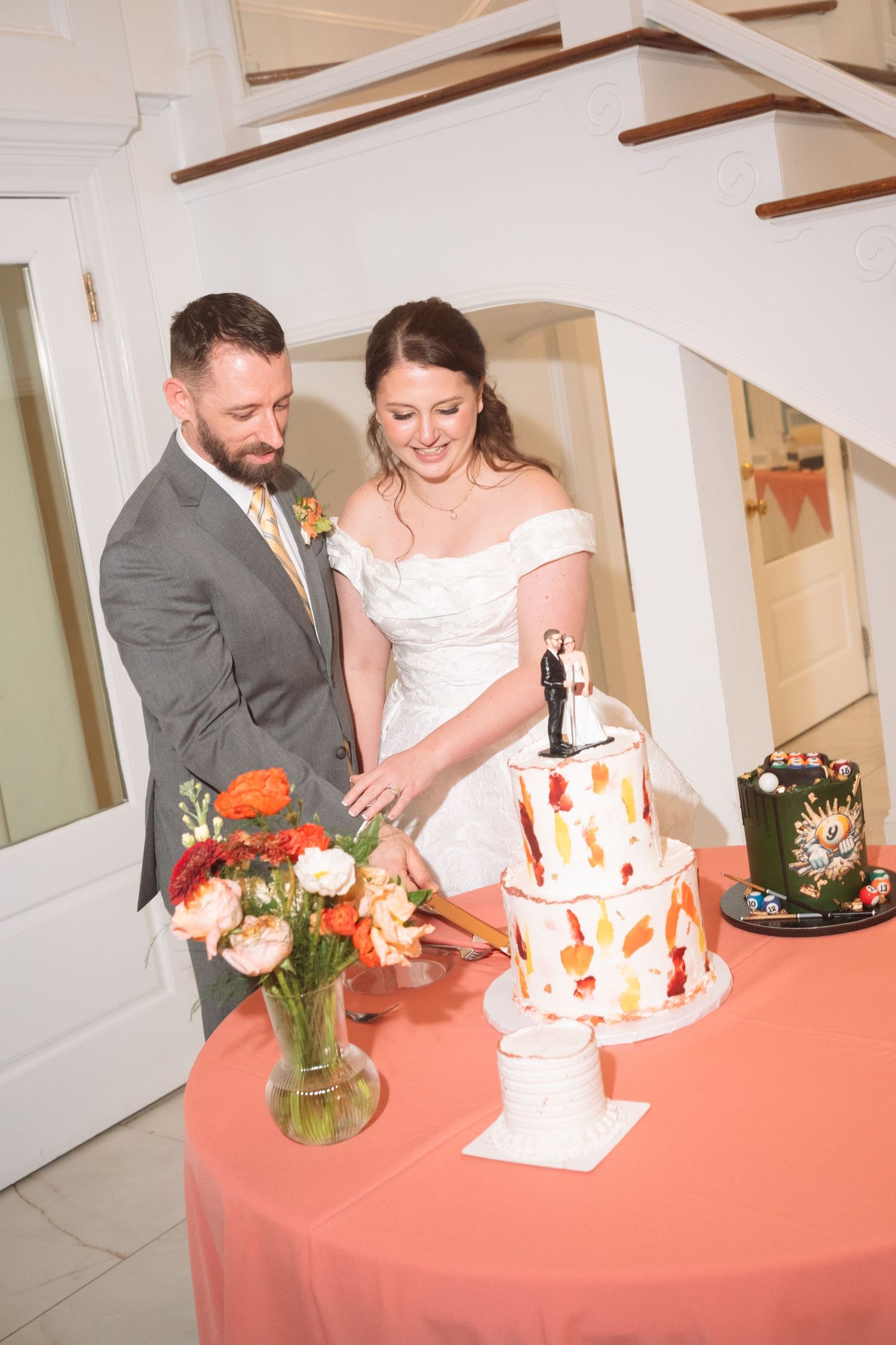 Bride and groom cutting their two-tier wedding cake with figurine topper at a reception table with orange floral arrangements.