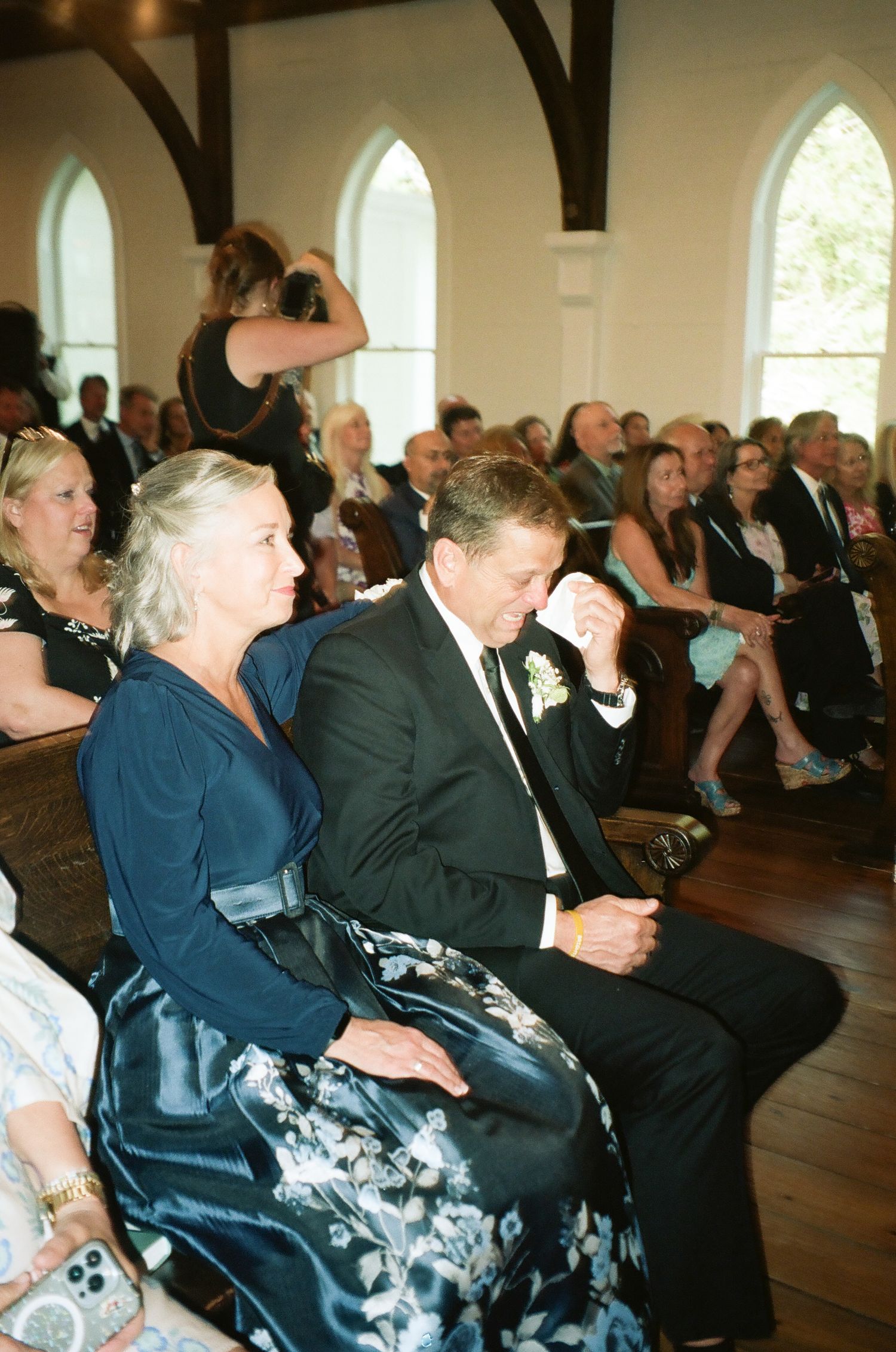 Emotional wedding guests seated in church pews, with man wiping tears during ceremony.