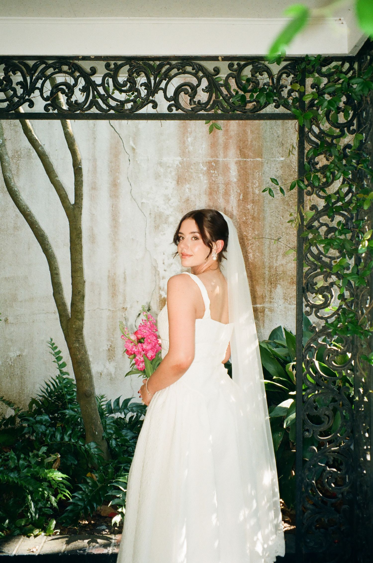 Bride in white gown holding pink bouquet stands before ornate iron gate with lush greenery backdrop.