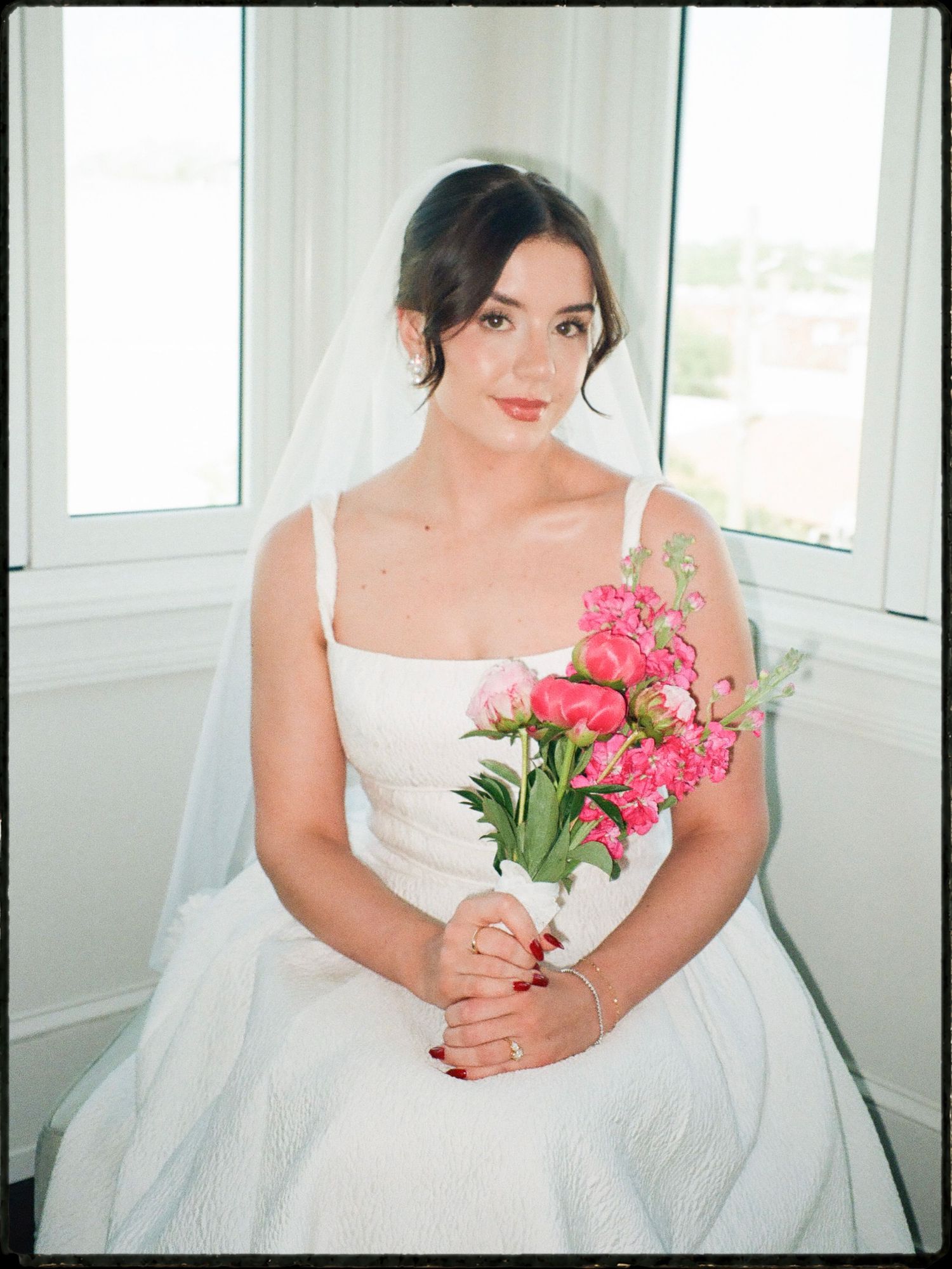 Elegant bride in white gown holds vibrant pink tulip bouquet while seated near bright window.