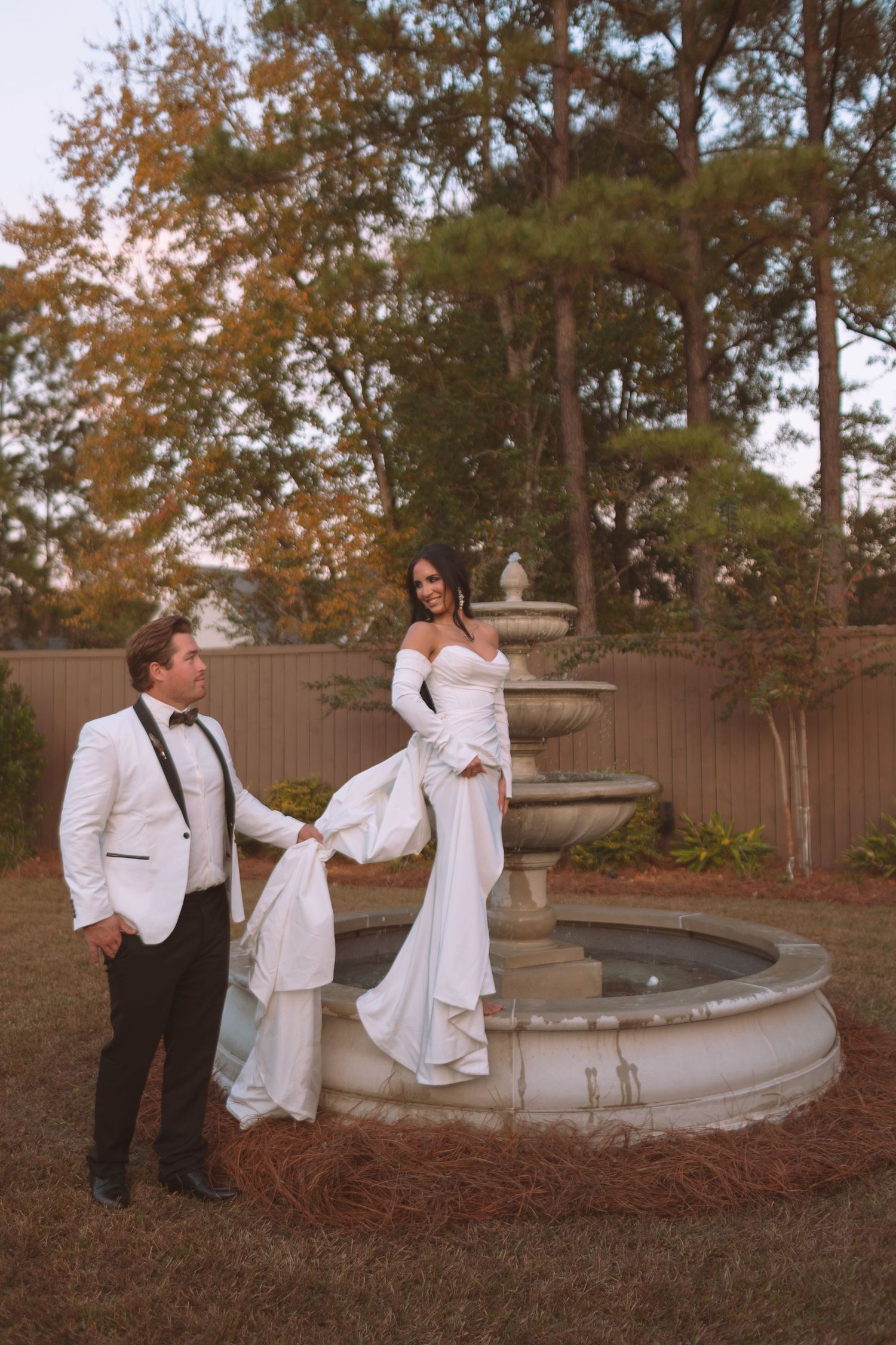 Bride and groom posing elegantly by a tiered stone fountain surrounded by autumn trees at dusk.