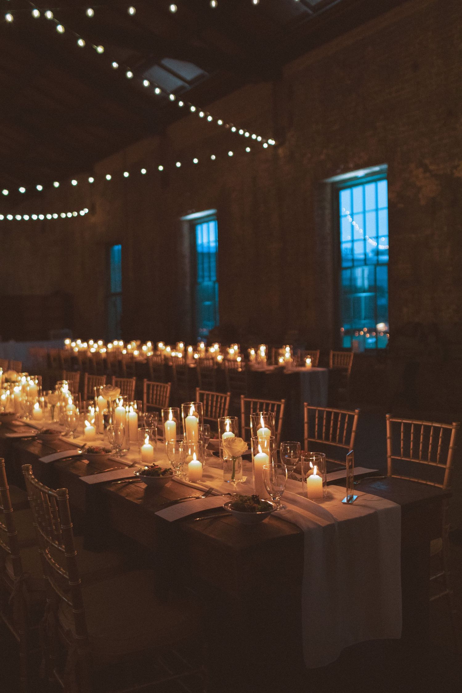 Elegant candlelit wedding reception tables in a rustic brick venue with string lights overhead.