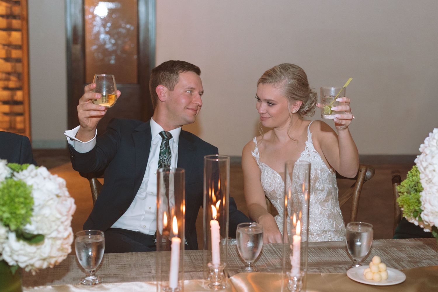 Bride and groom raising glasses in toast at candlelit wedding reception sweetheart table.