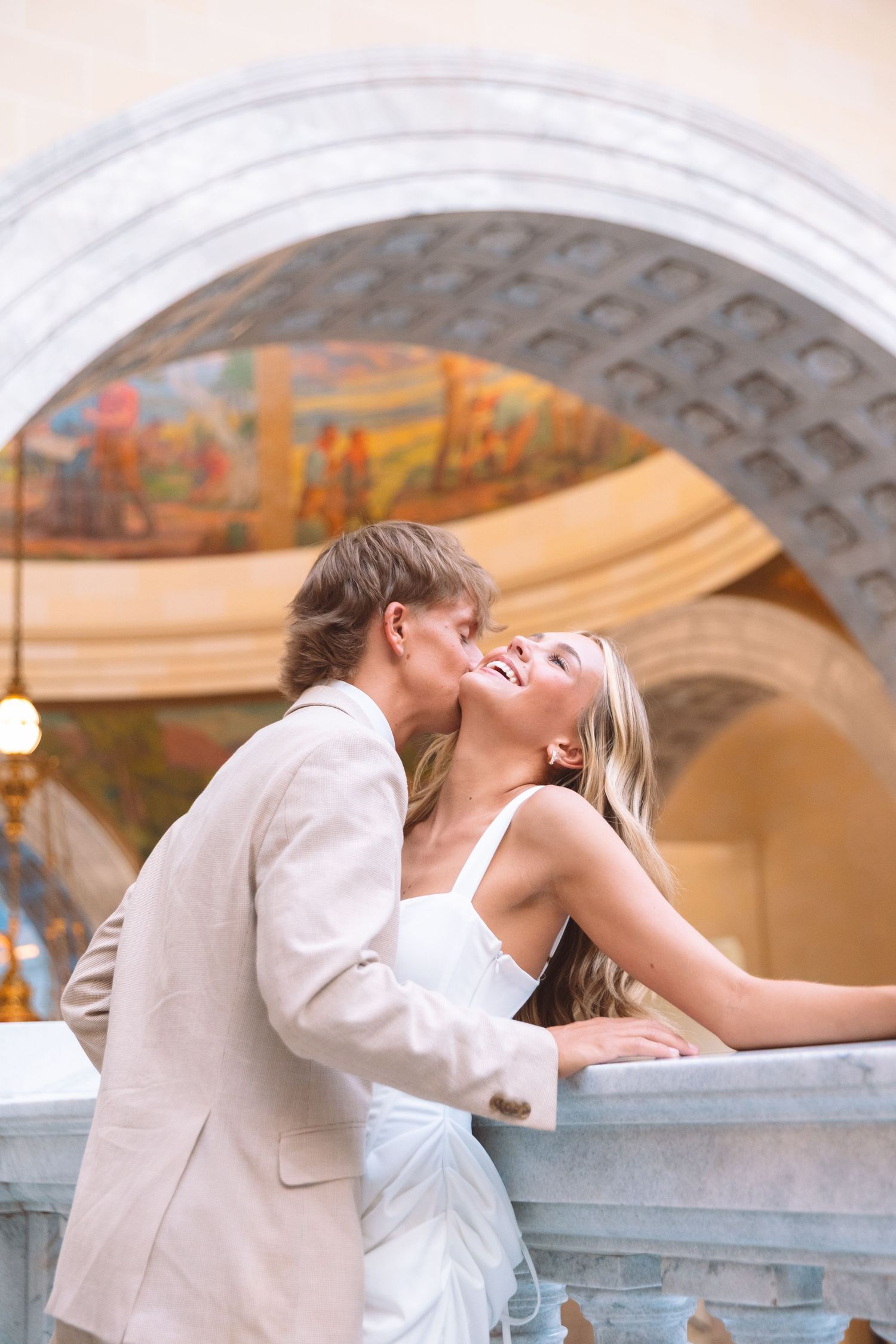 A groom in a beige suit kisses his smiling bride in a white dress on a elegant balcony with arched architecture.