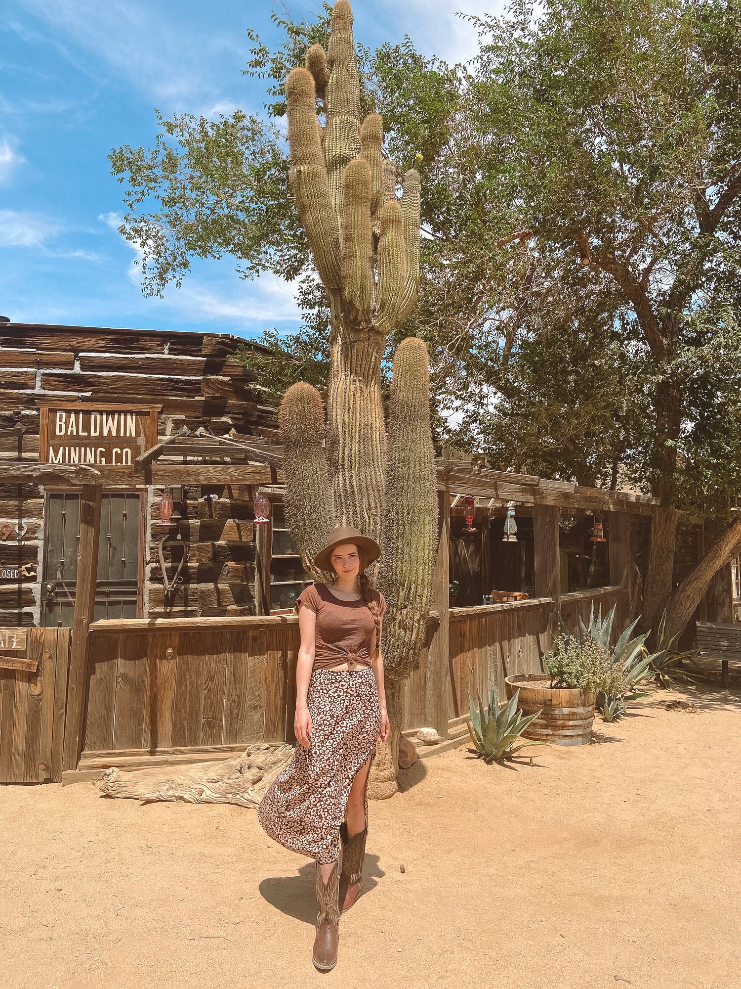 Woman in floral skirt and cowboy hat poses beside tall saguaro cactus at Baldwin Mining Co. desert attraction.