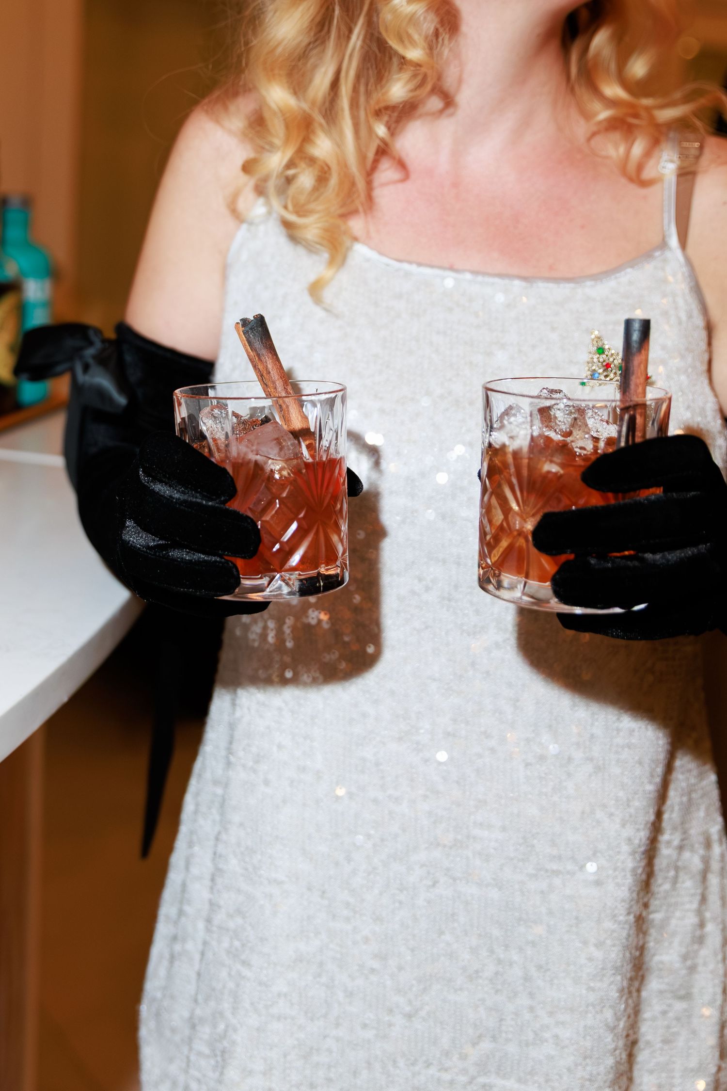 A woman in a silver sequin dress wearing black gloves holds two cocktails with cinnamon sticks.