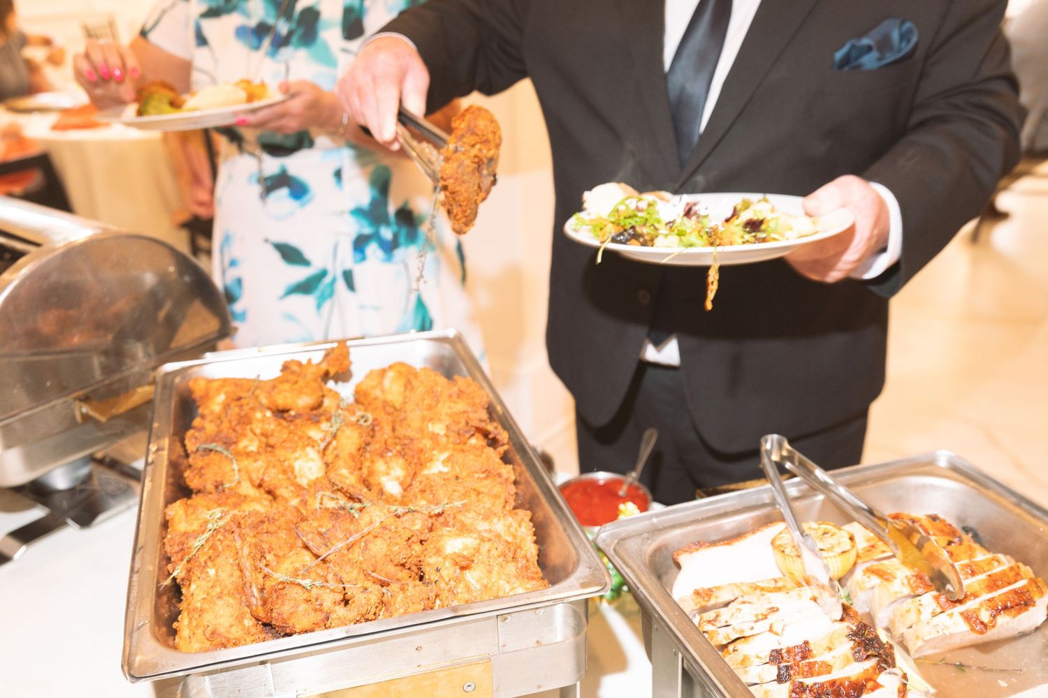 Man in suit serving fried chicken and stuffed bread rolls at a catered buffet.