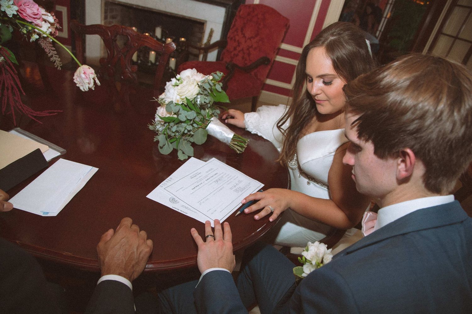 Bride and groom sign wedding certificate at ornate wooden table with floral bouquet nearby.