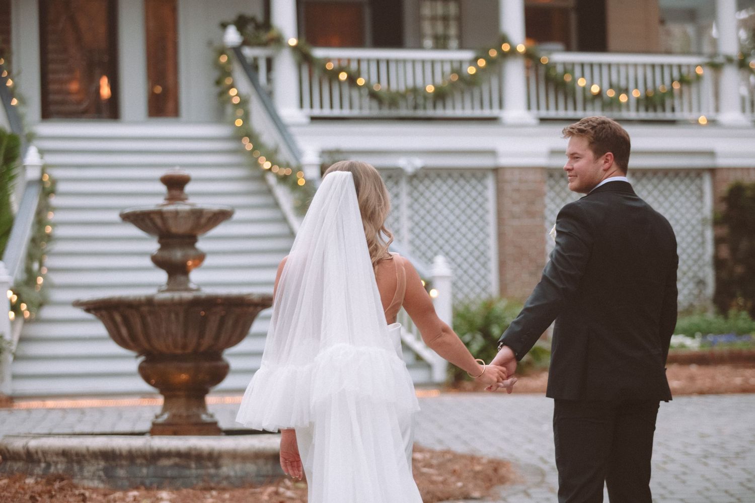 Bride and groom hold hands walking toward white colonial estate adorned with string lights.