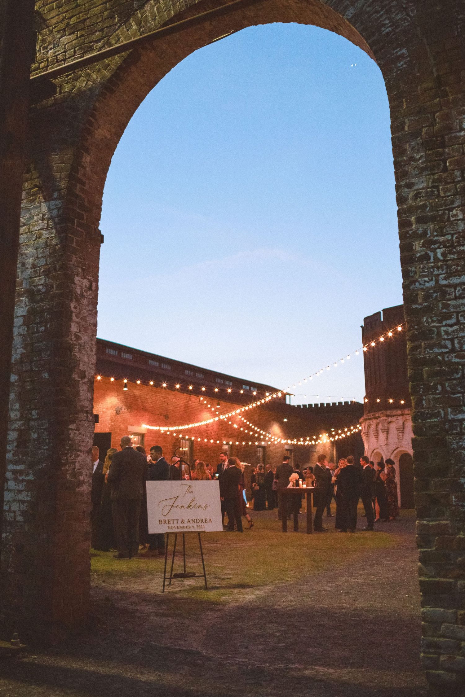 Wedding guests mingling in a historic brick courtyard venue at dusk with warm string lights overhead.