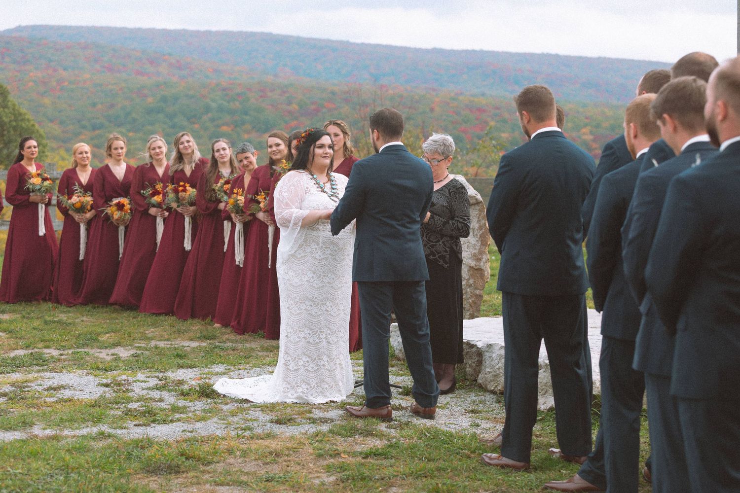 Outdoor fall wedding ceremony with bridesmaids in burgundy and groomsmen in navy on hillside.