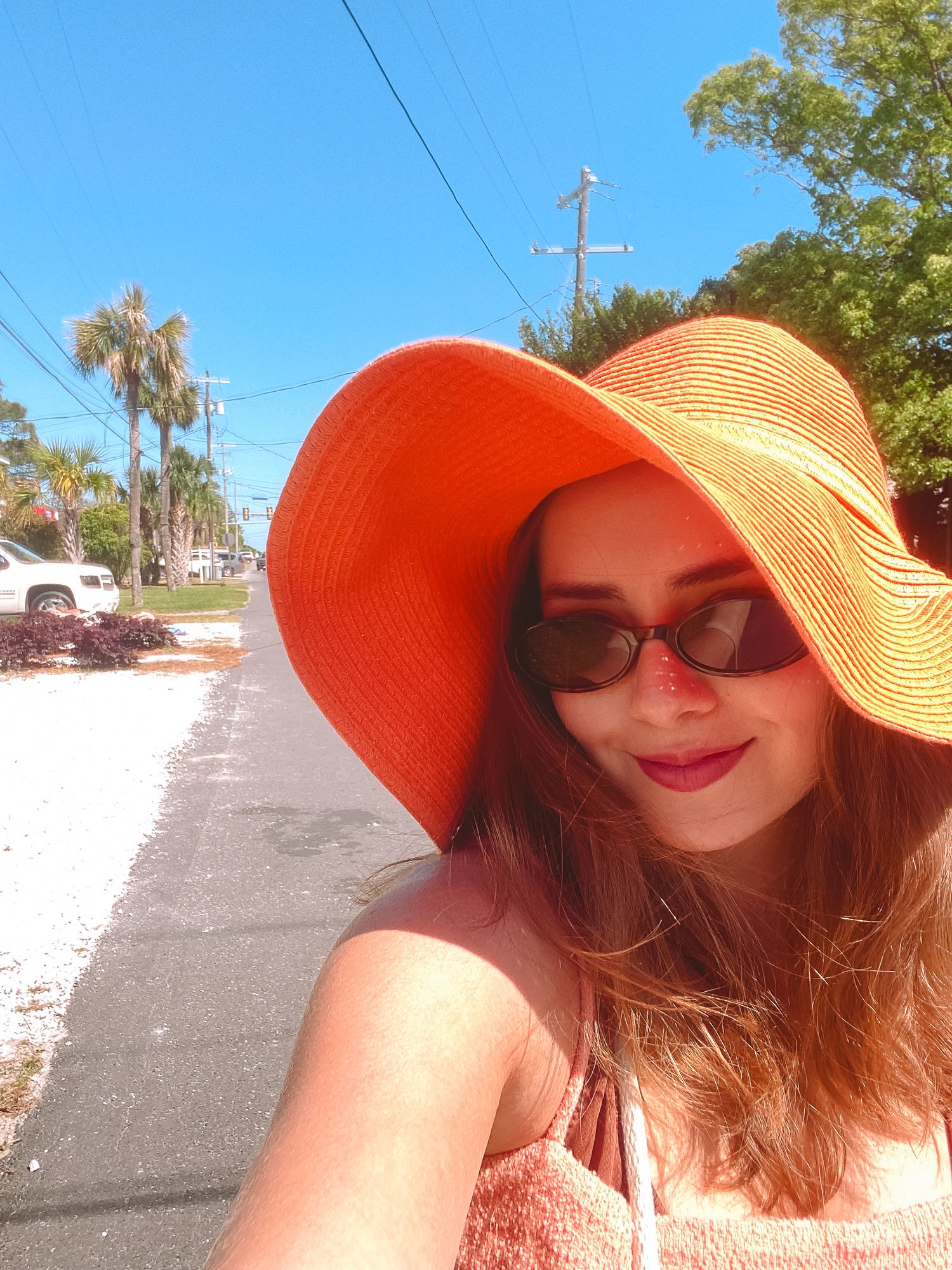 Woman taking a sunny selfie wearing a large orange floppy sun hat and sunglasses on a tree-lined street.