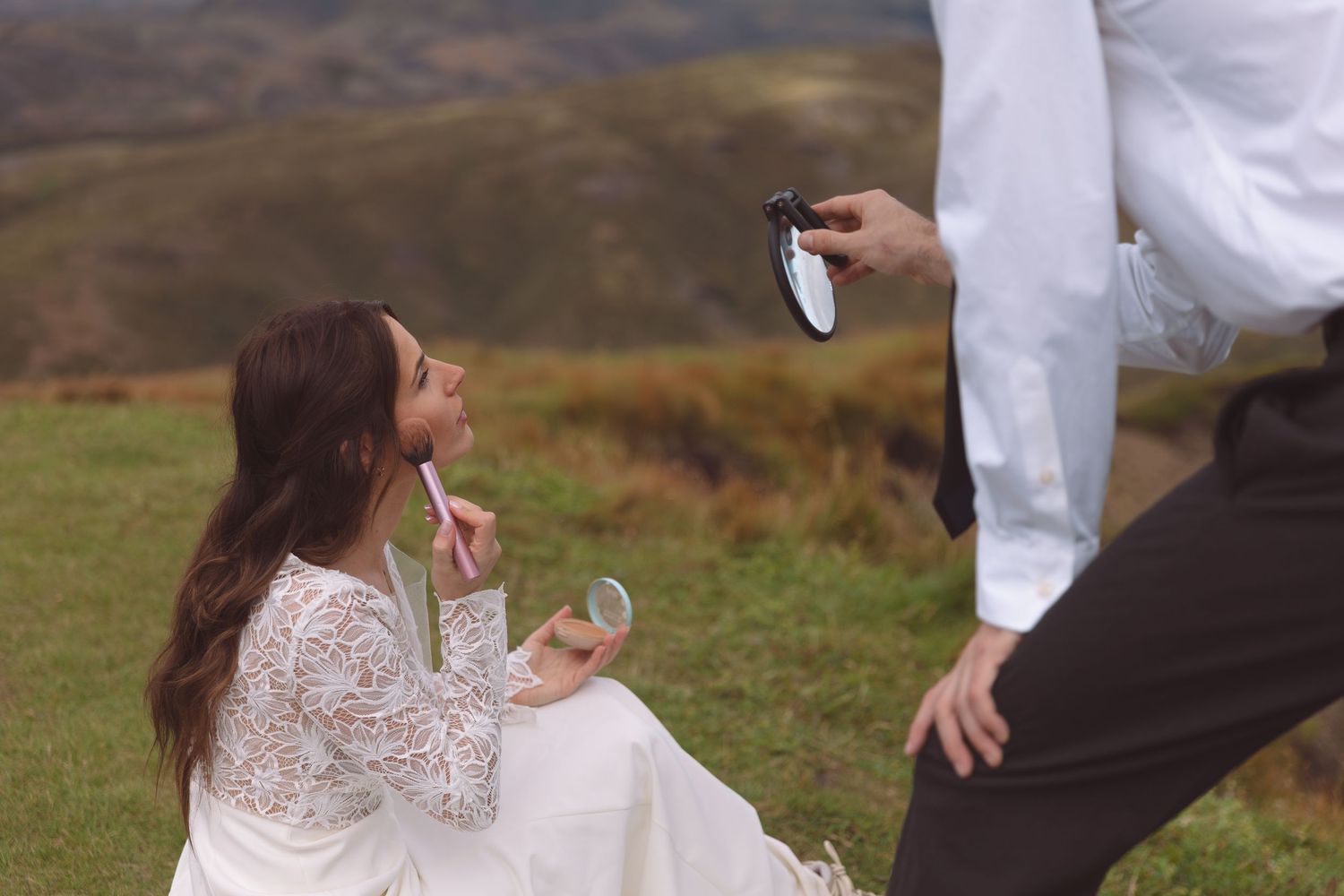 Bride applies lipstick using small mirror held by groom during outdoor mountain wedding photography session.