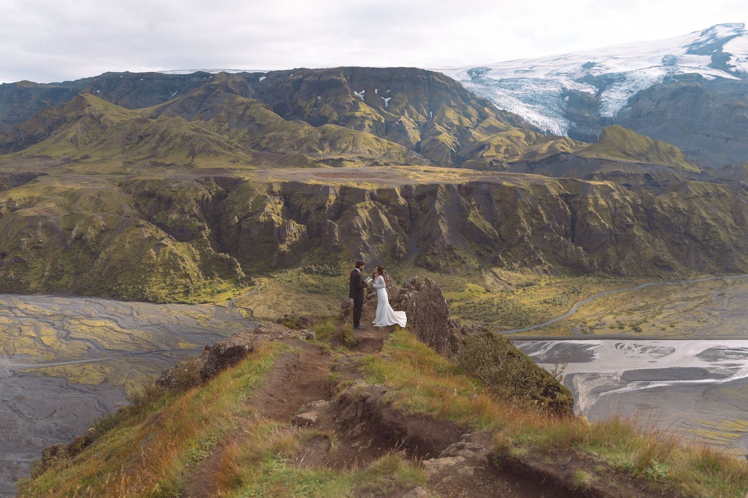 Bride and groom overlooking dramatic Icelandic river valley and glacier from hilltop viewpoint.