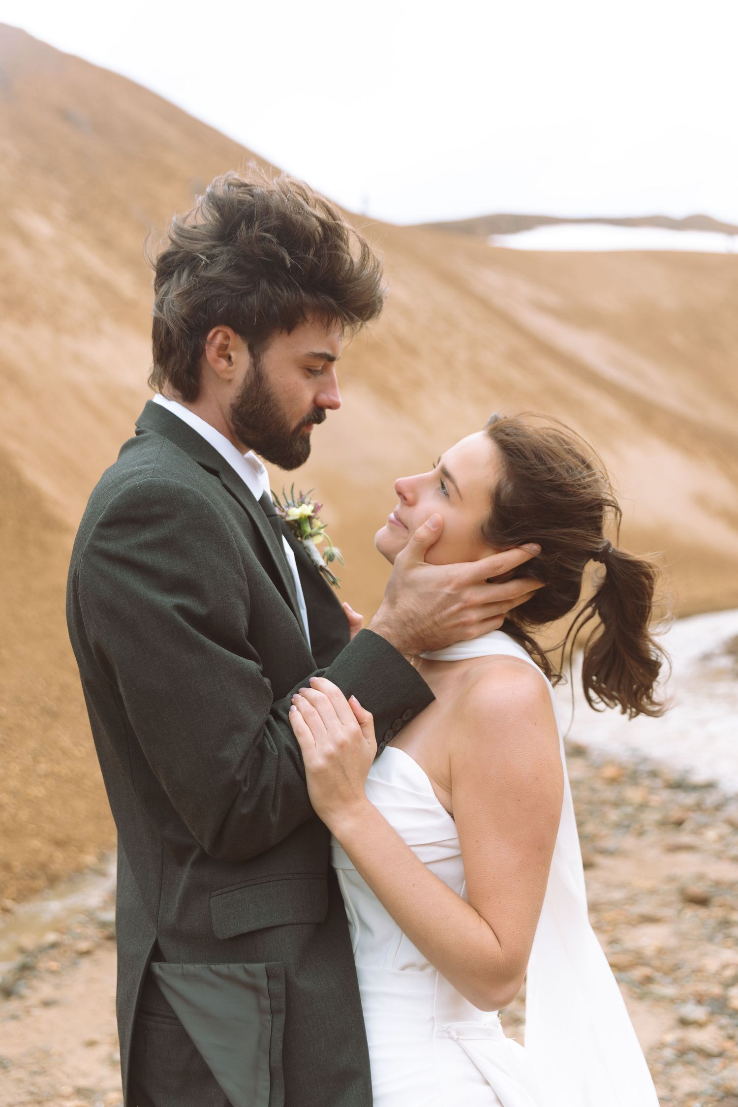 Couple shares an intimate moment, groom gently holding bride's face as they gaze at each other in mountain landscape.