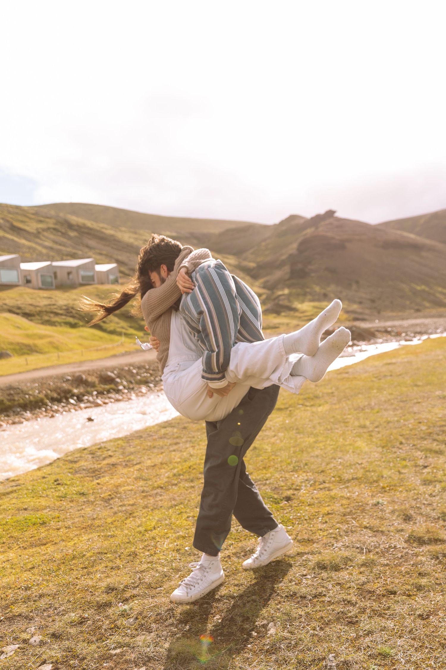 Man carrying woman in his arms in bright sunlight on a green Icelandic hillside near a river.
