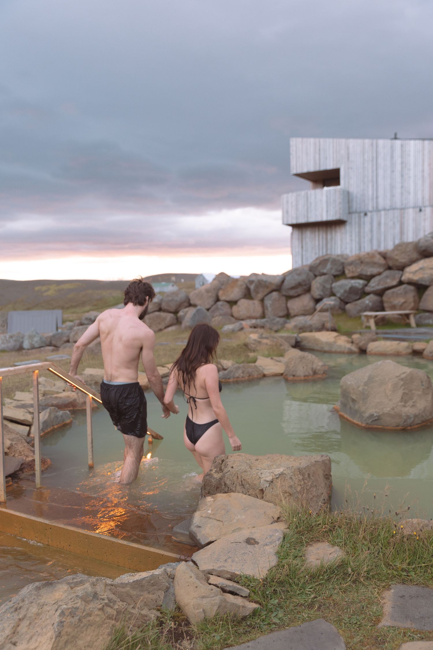 Couple holding hands walking into a natural geothermal pool at sunset with modern stone building in Iceland.
