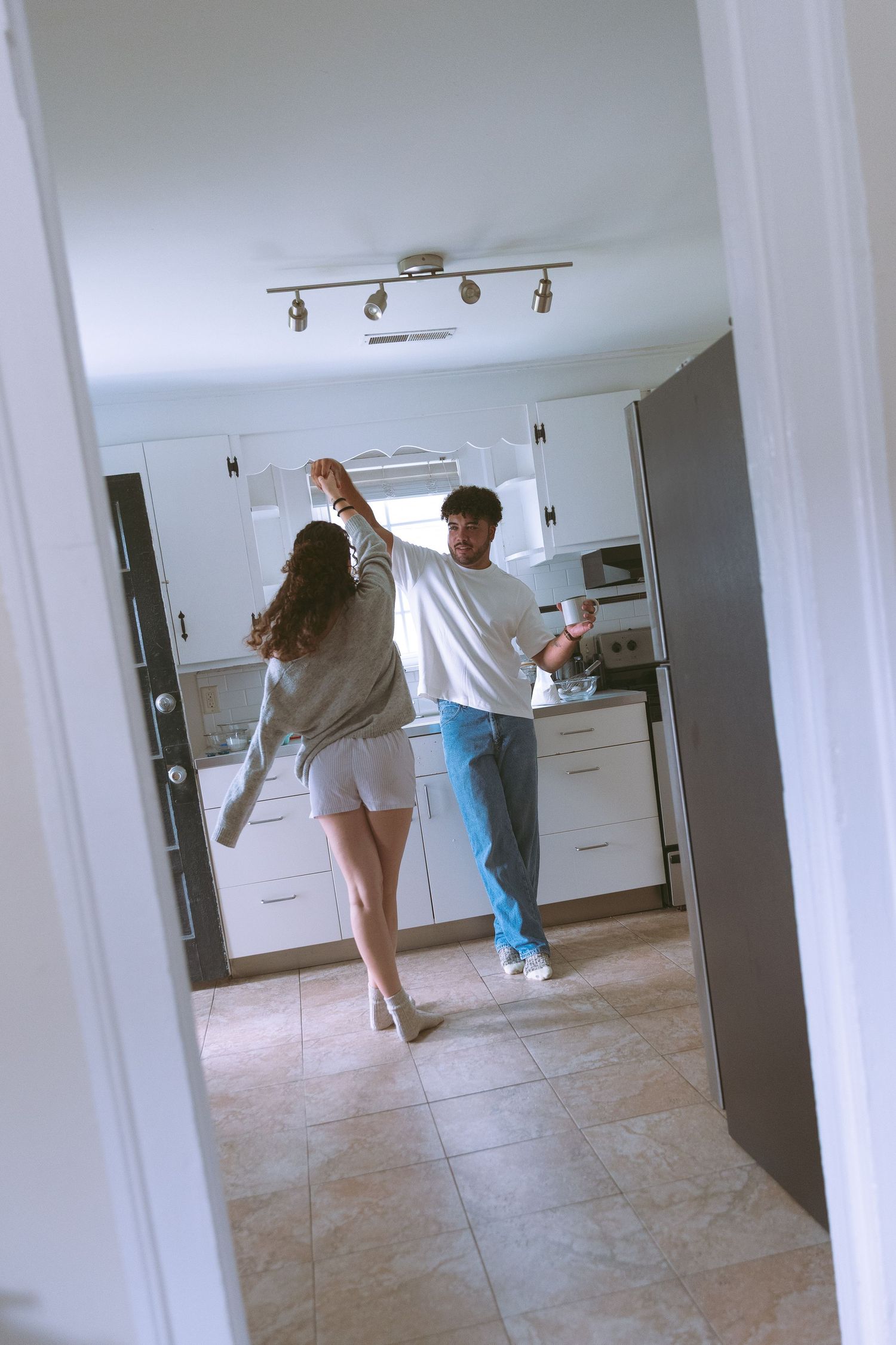 Couple dancing together in a bright white kitchen, viewed through a doorway, man spinning woman around.