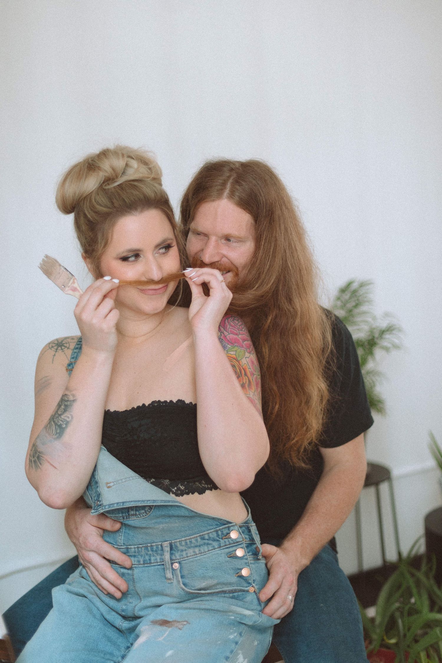 Couple playfully using a paintbrush as a fake mustache while smiling indoors.