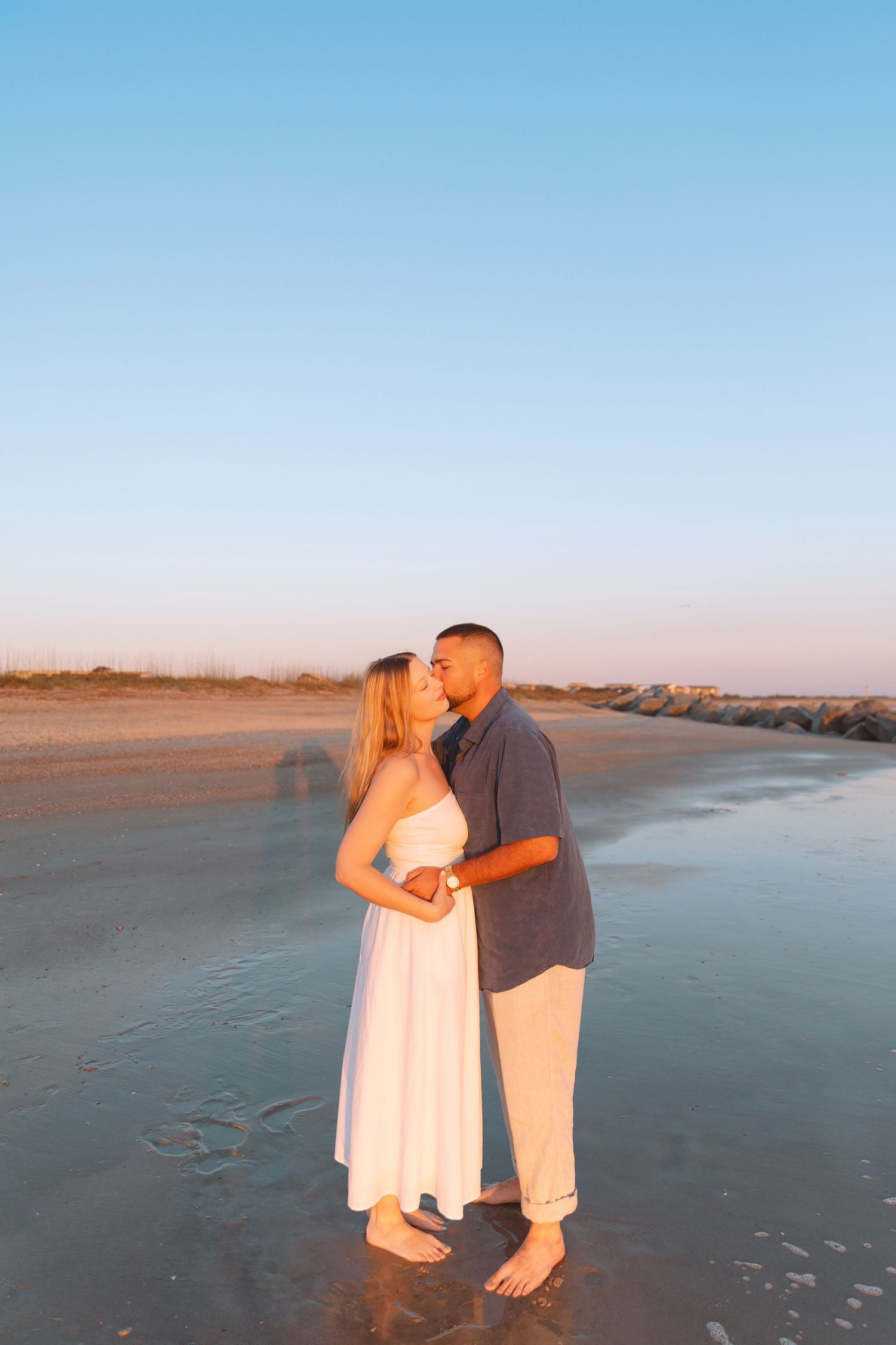 Couple sharing a romantic kiss on a sandy beach at golden hour, woman in white dress, man in gray shirt.