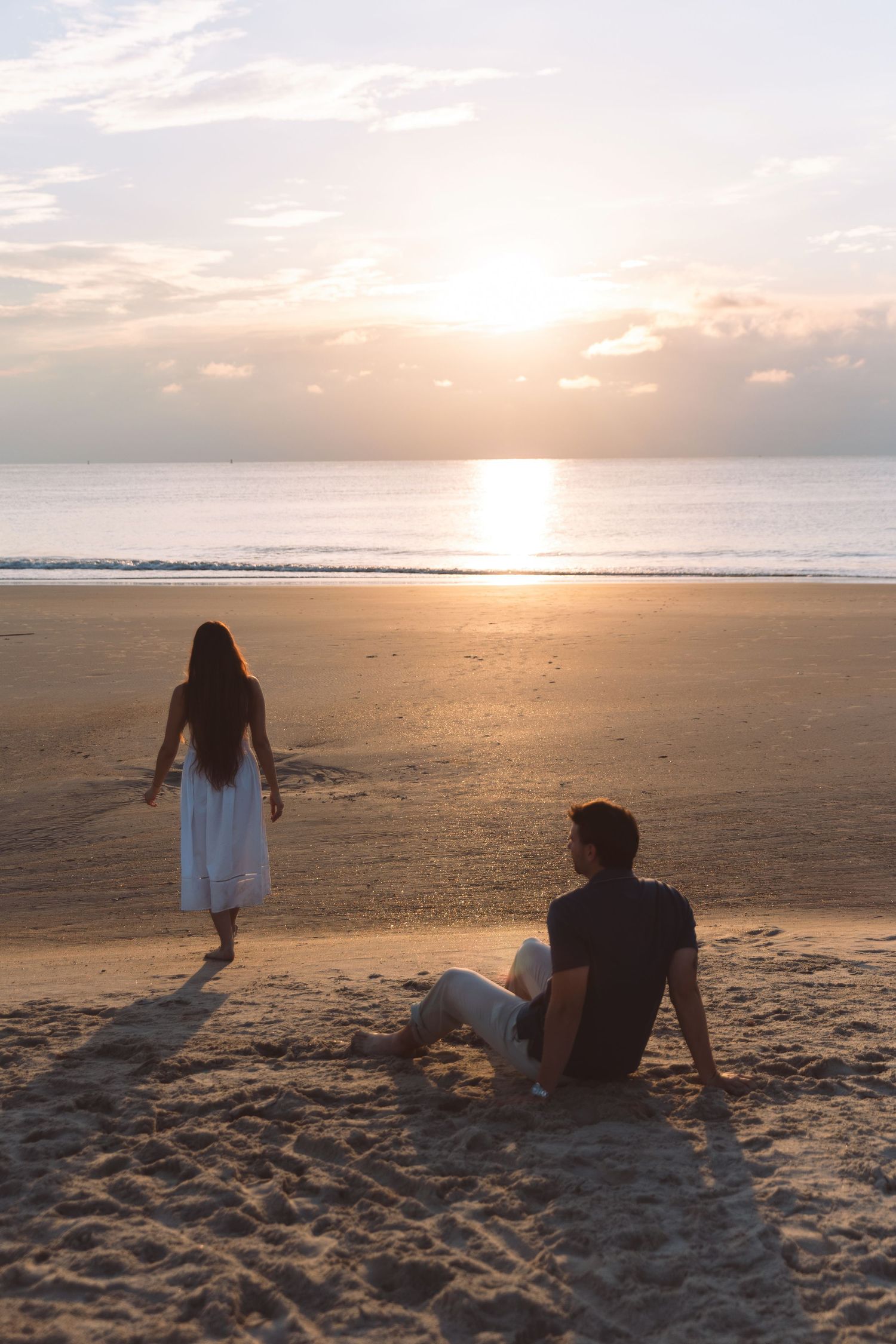 Couple on sandy beach at sunset, woman in white dress walking toward ocean while man sits watching the golden horizon.