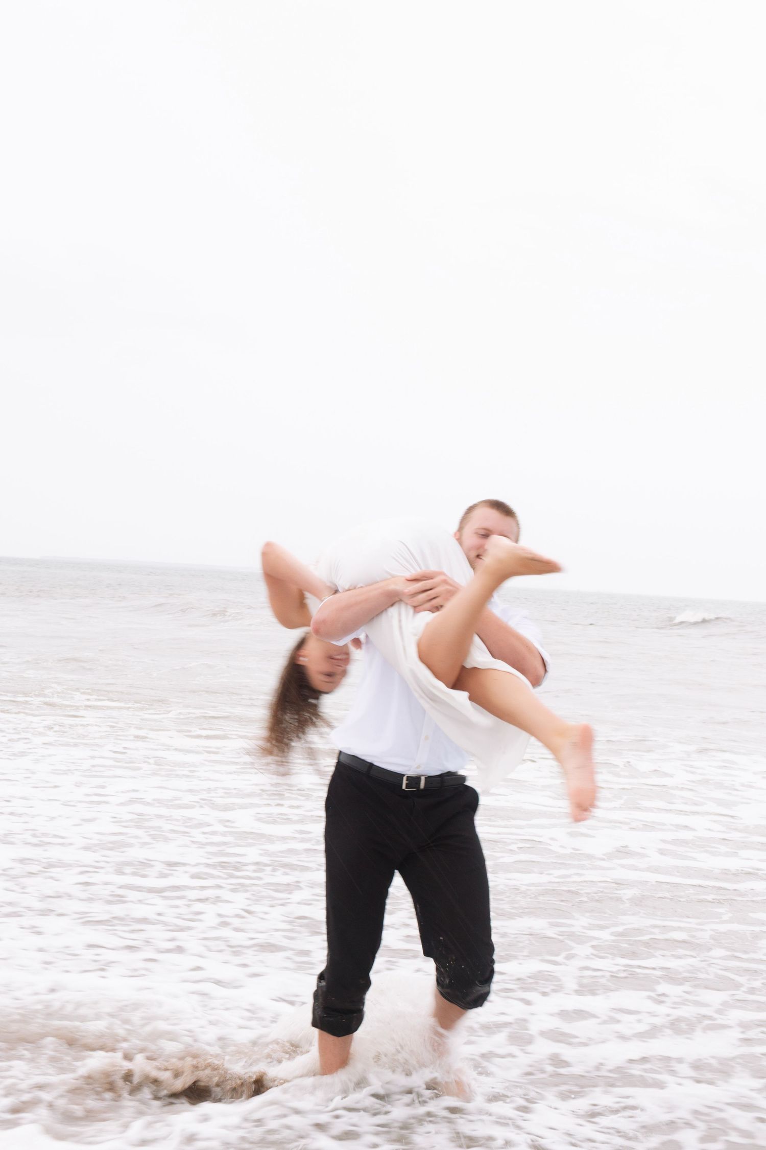 Man playfully dipping woman in ocean waves on the beach during a romantic photo shoot.